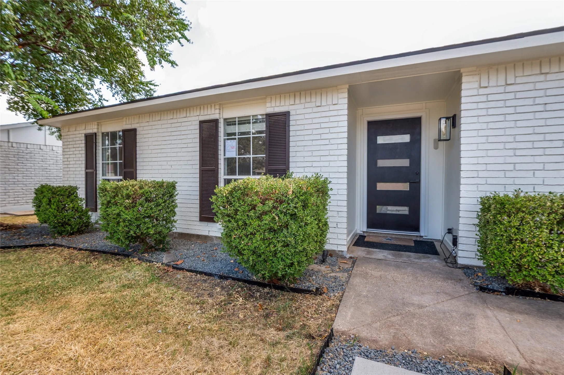 White brick exterior, contemporary front door with horizontal glass inserts, black exterior lighting fixture, dark shutters, and landscaped beds with hedging
