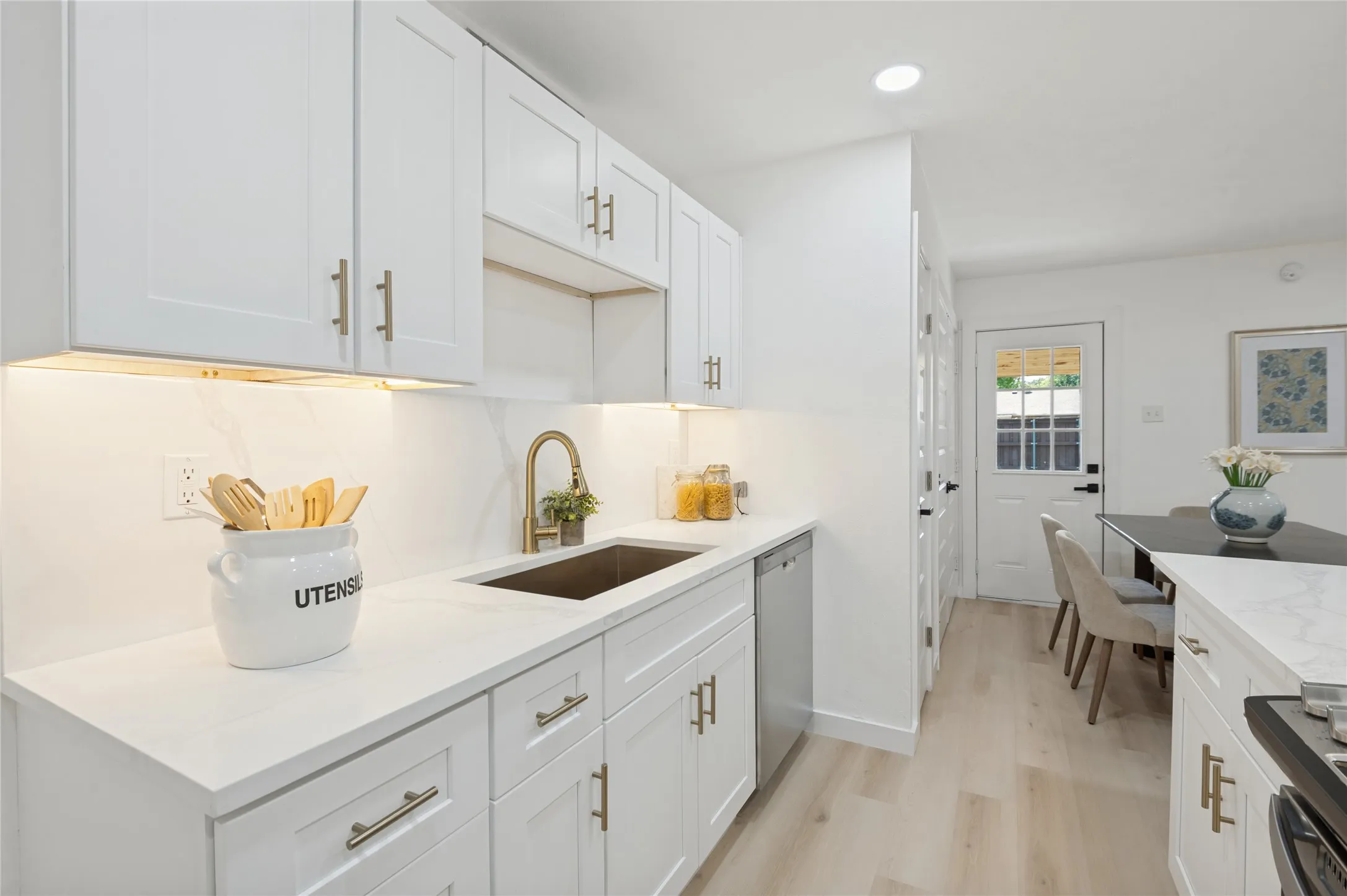 White cabinetry with gold-finish hardware, light-toned wood-finish flooring, undermount sink with gold-finish faucet, white countertops, and stainless-steel dishwasher