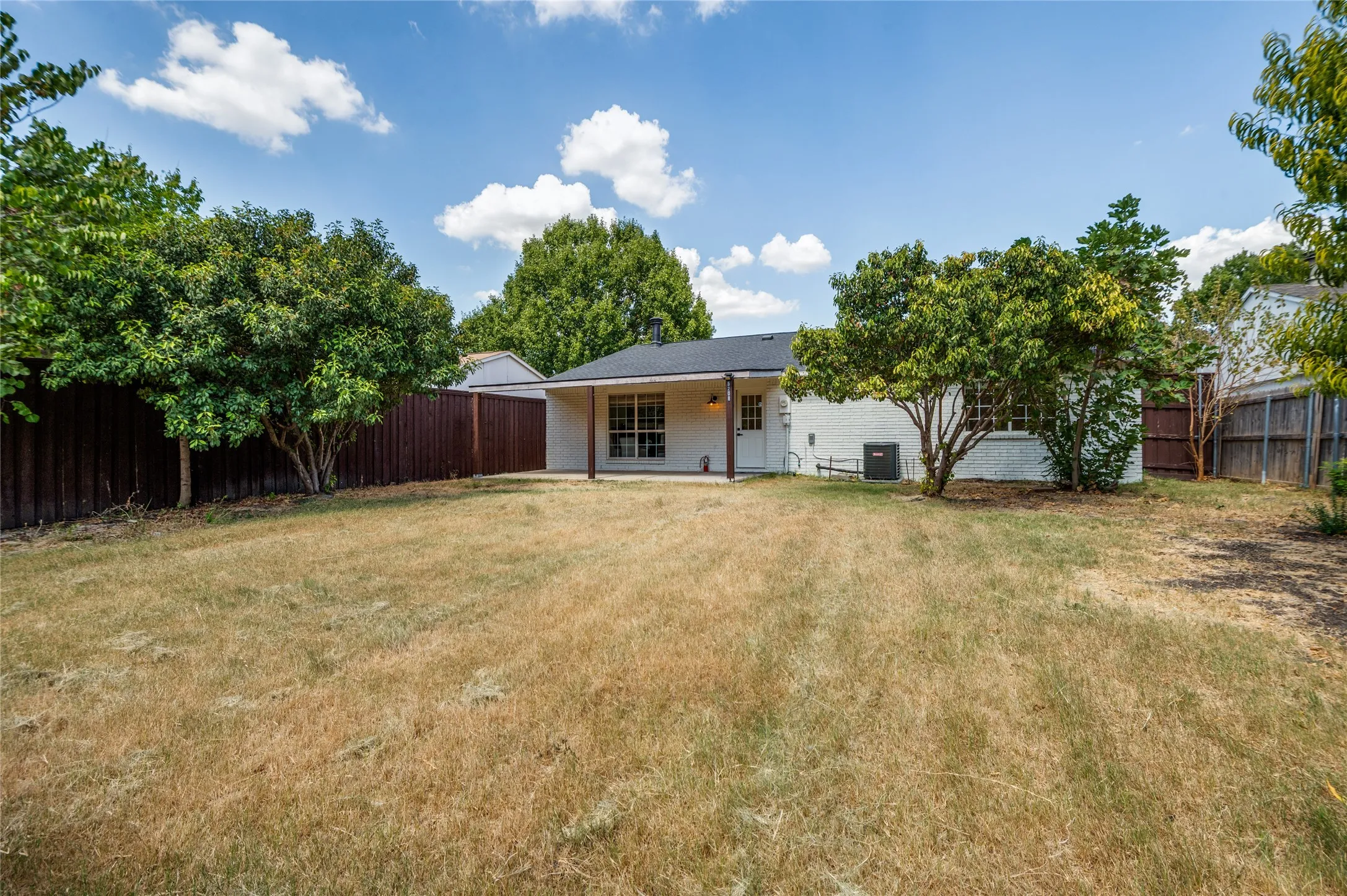 Expansive backyard featuring a partial privacy fence, mature trees, and a white brick exterior