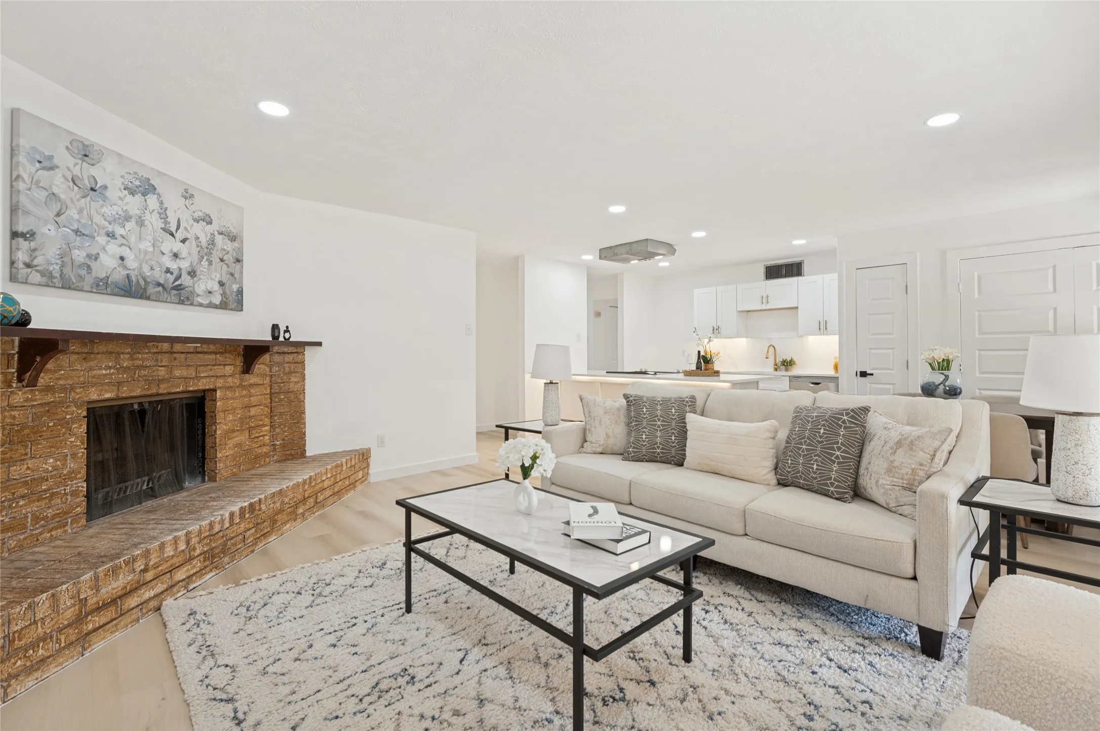 Recessed lighting, wood-finish flooring, brick fireplace with wood mantel, kitchen with white cabinetry, and a gold-finish faucet