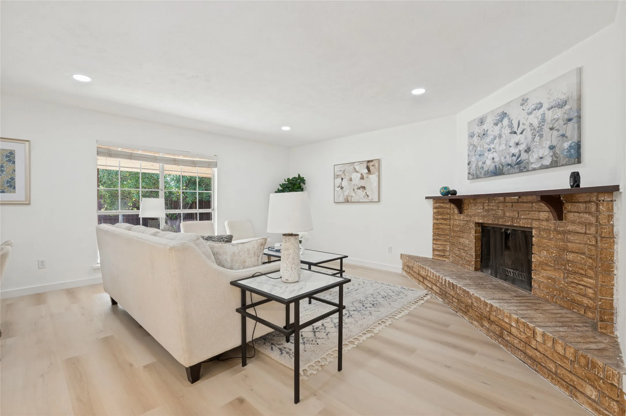 Spacious living area featuring light wood-finish flooring, a prominent brick fireplace with a wood mantle, and recessed lighting