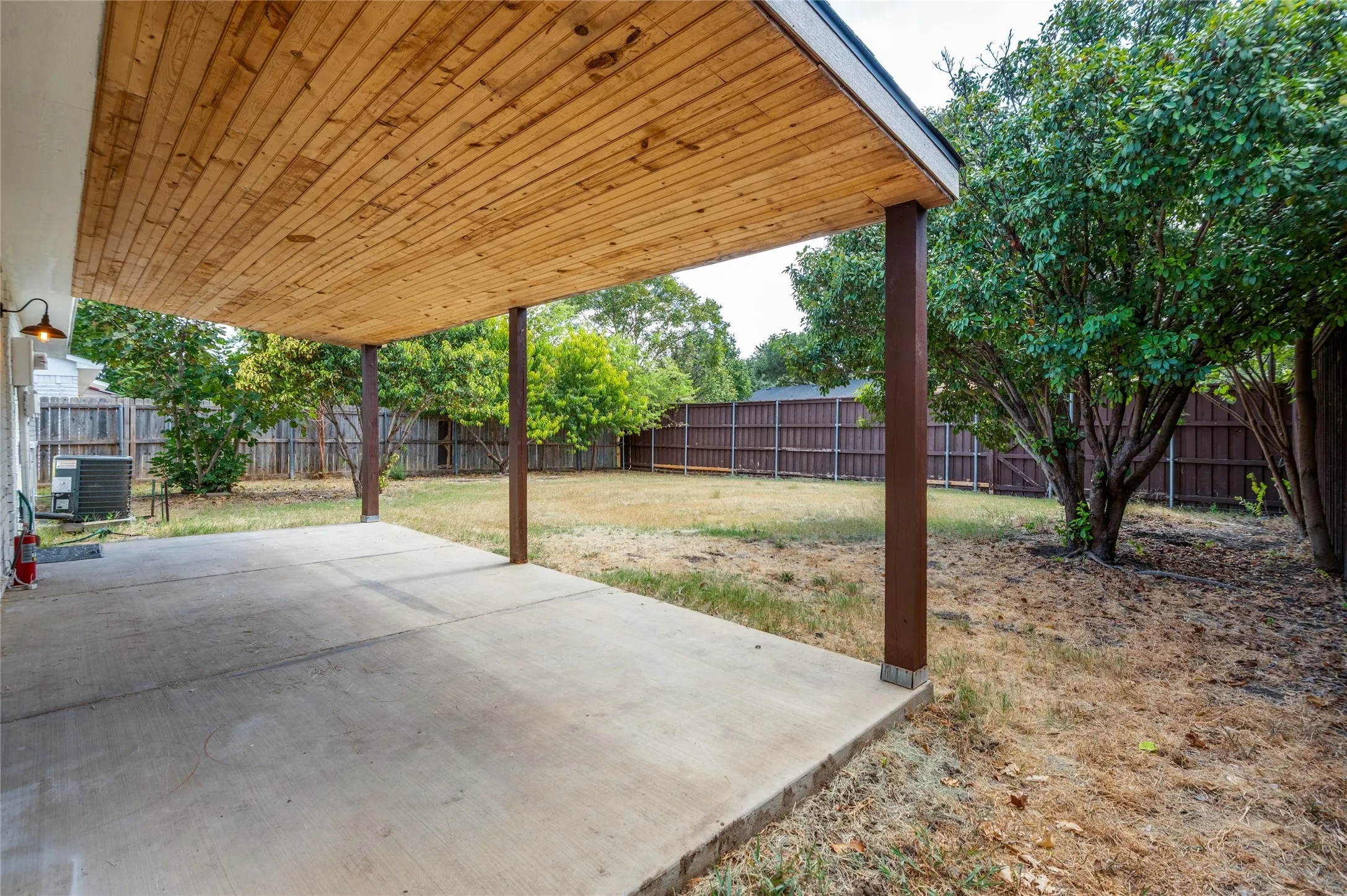 Covered concrete patio with a wood-finish ceiling, supported by posts, opening to a fenced yard with mature trees