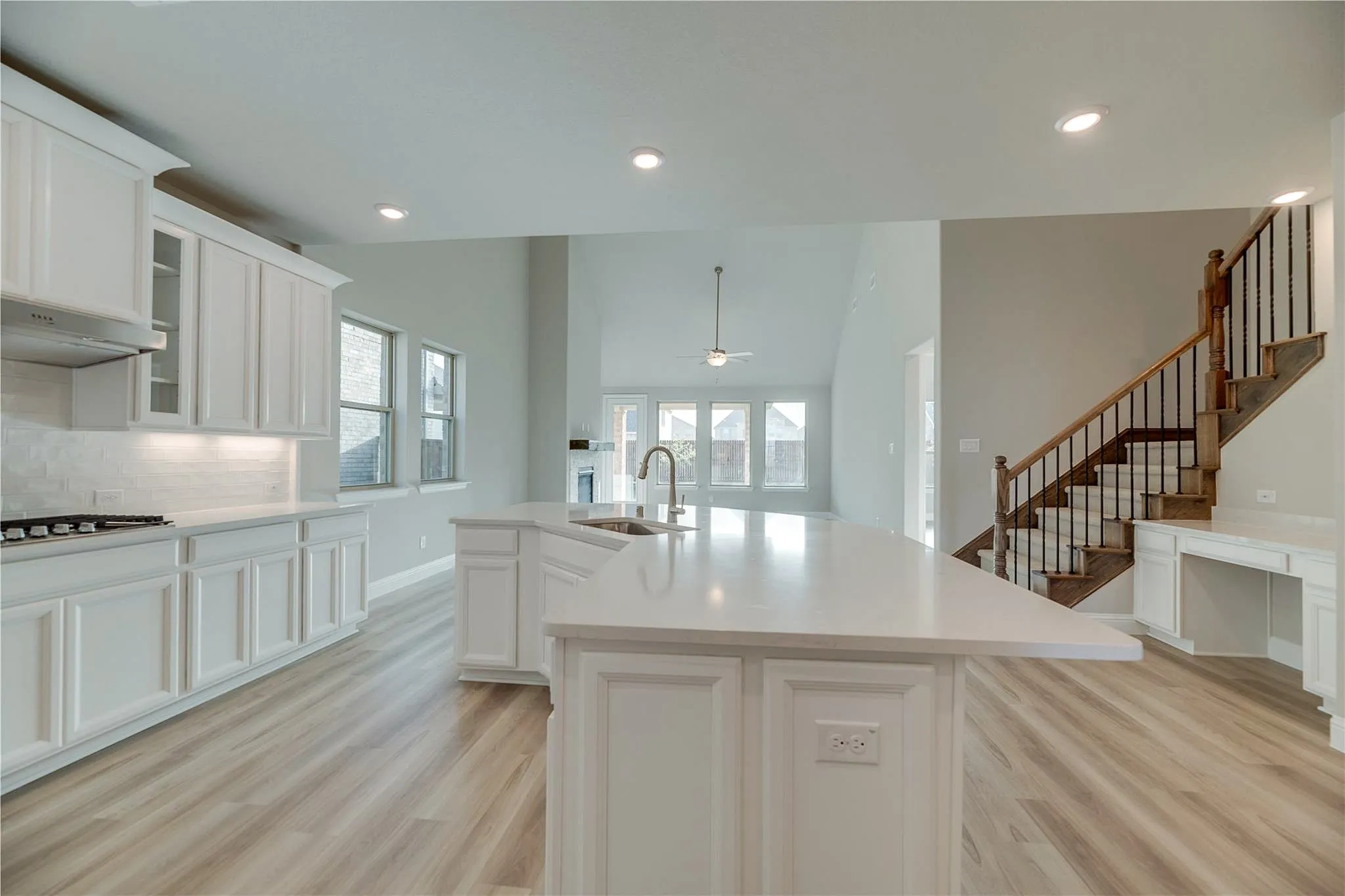 Kitchen island with a built-in sink and white countertops, white cabinetry with under-cabinet lighting, wood-finish flooring, a staircase with wood railing and dark metal spindles, and a built-in desk area