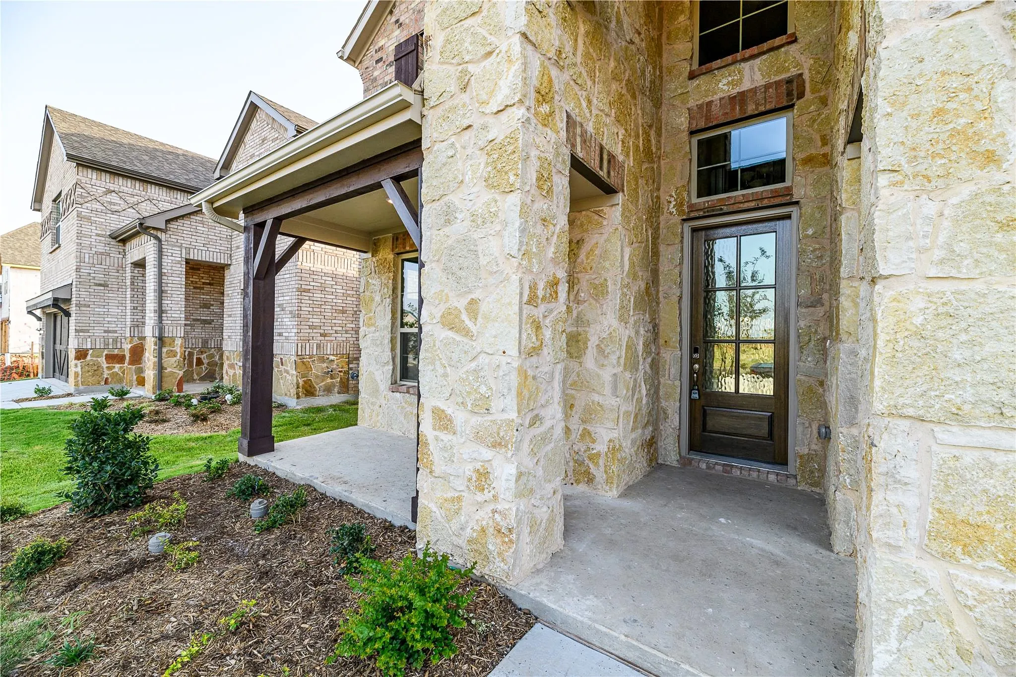Stone and brick exterior with a covered entry, dark wood-finish front door with glass panels, and decorative landscaping