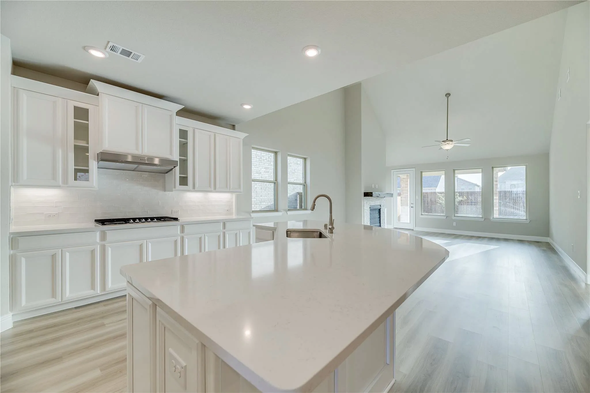 Kitchen island with a sink and gooseneck faucet, white shaker cabinetry, stainless steel range hood, light-toned wood-finish flooring, and a vaulted ceiling