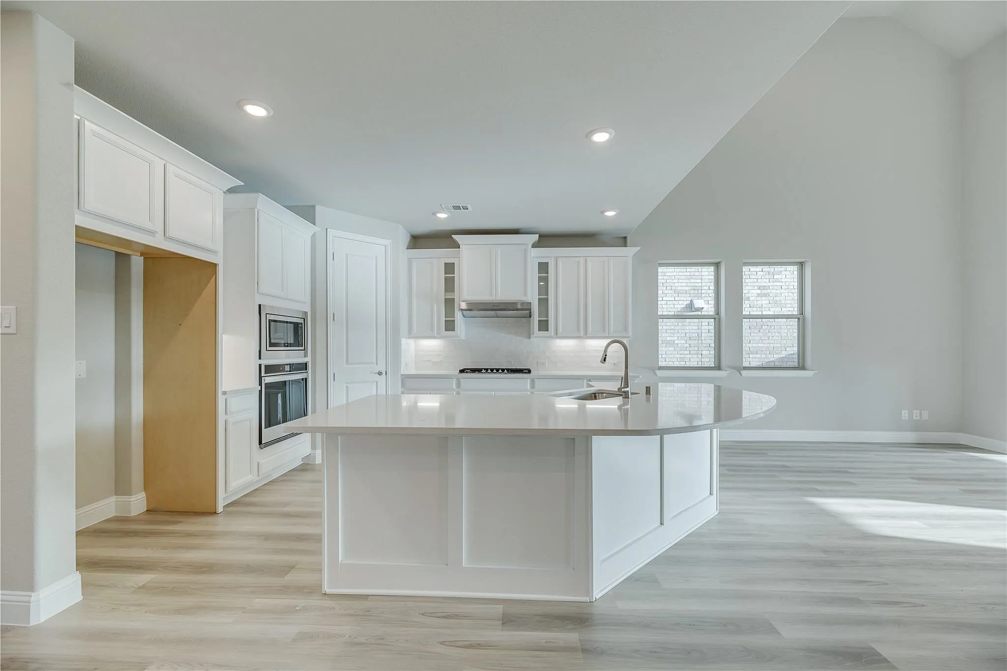 Open-concept kitchen with a curved island, white shaker cabinetry, built-in double ovens, and wood-finish flooring