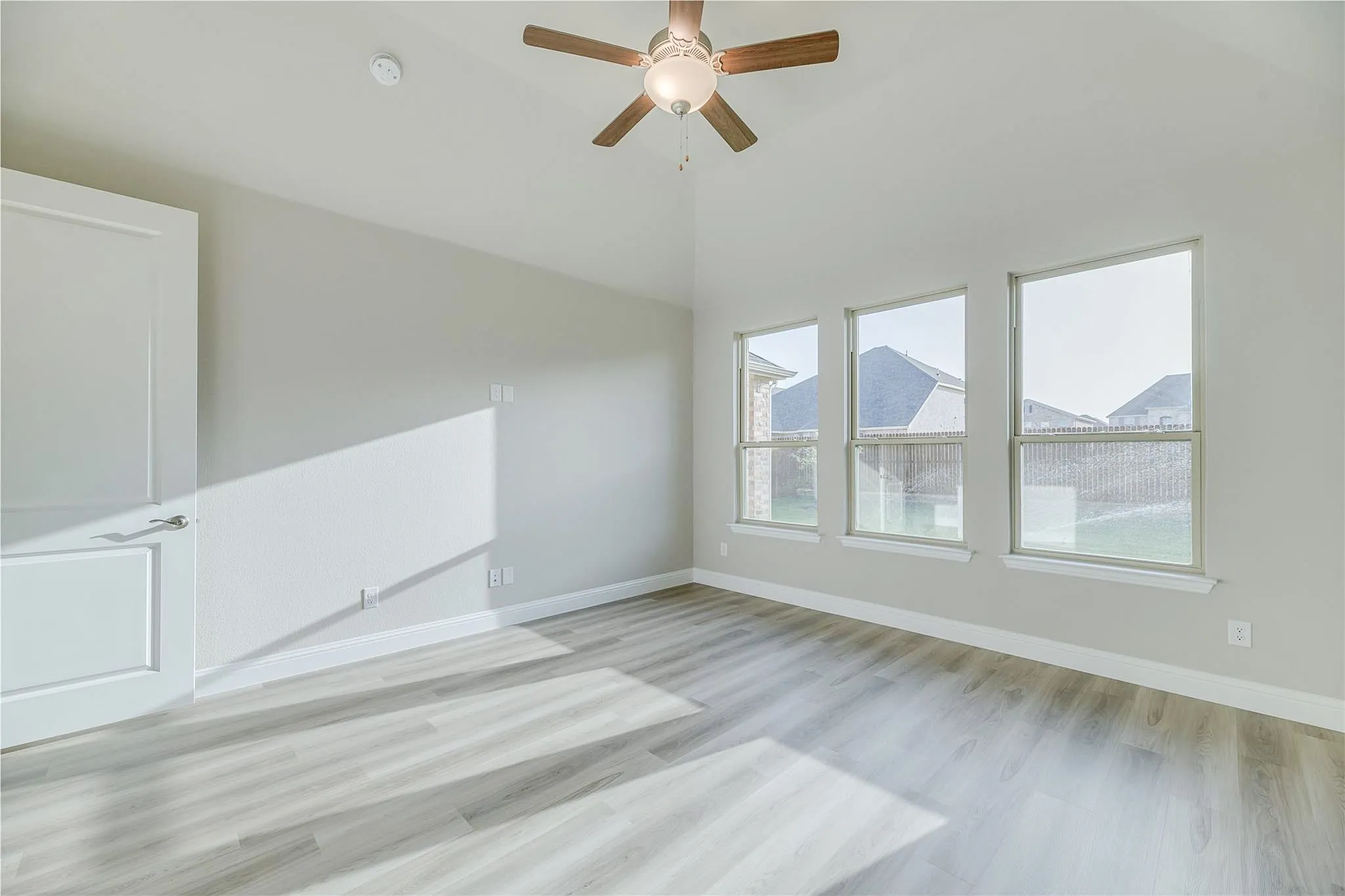 Vaulted ceiling with a ceiling fan, three vertical windows, white paneled door, light-toned wood-finish flooring