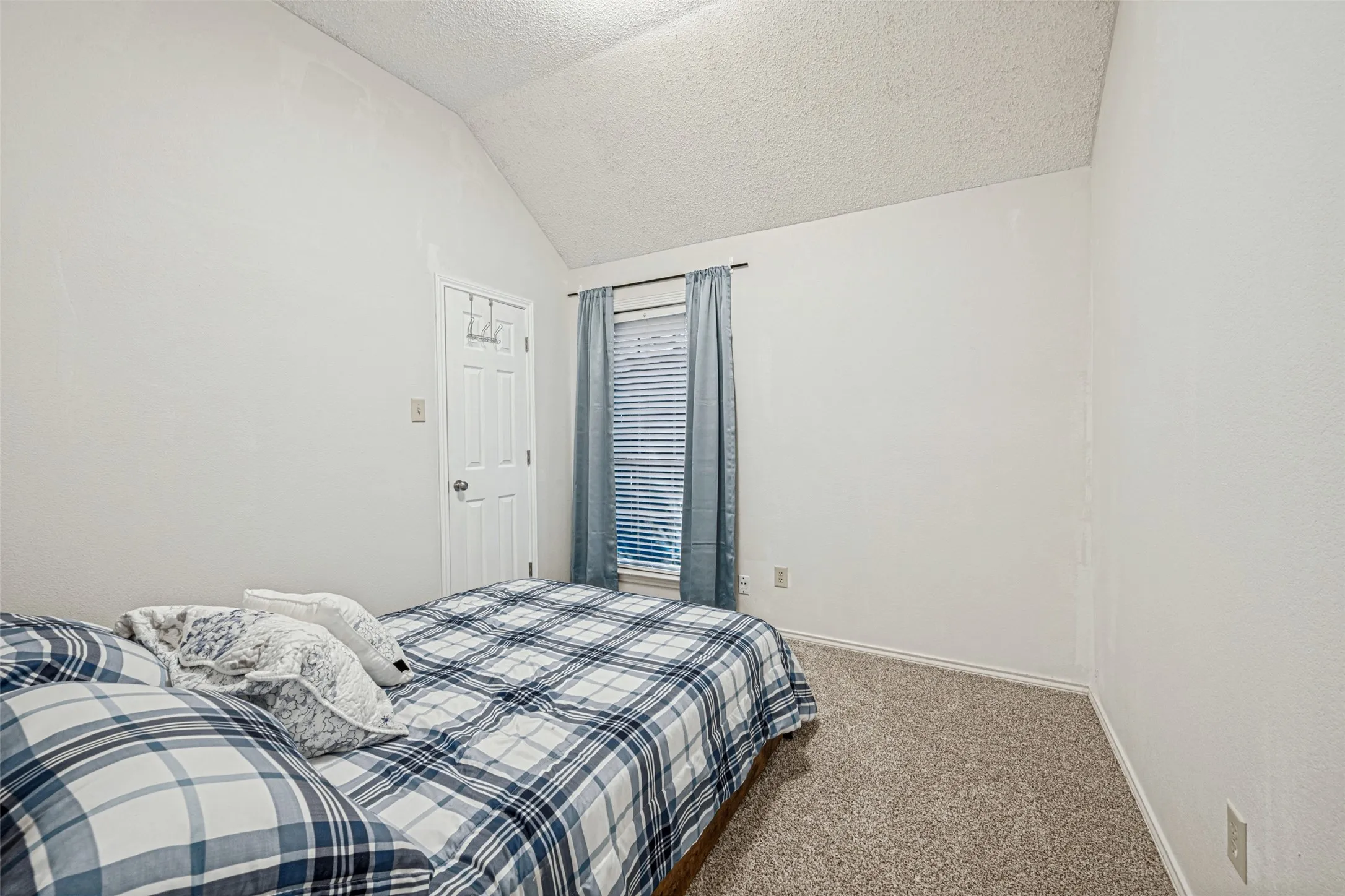 Vaulted ceiling, textured walls, window with blinds, carpeted flooring, and a paneled interior door