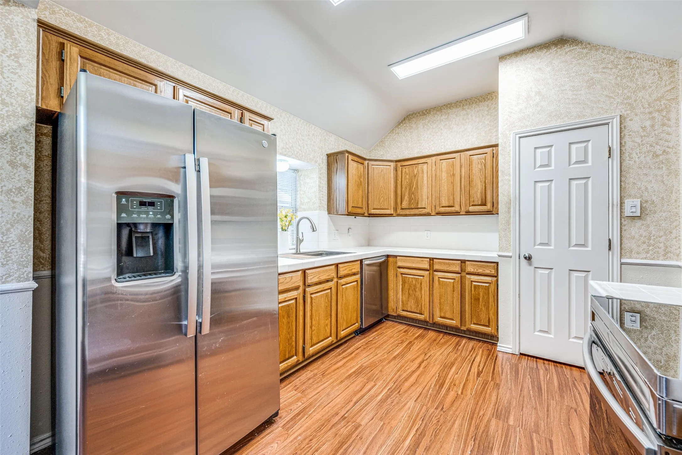 Kitchen featuring wood-finish flooring, natural wood cabinetry, stainless steel refrigerator, stainless steel dishwasher, and a vaulted ceiling
