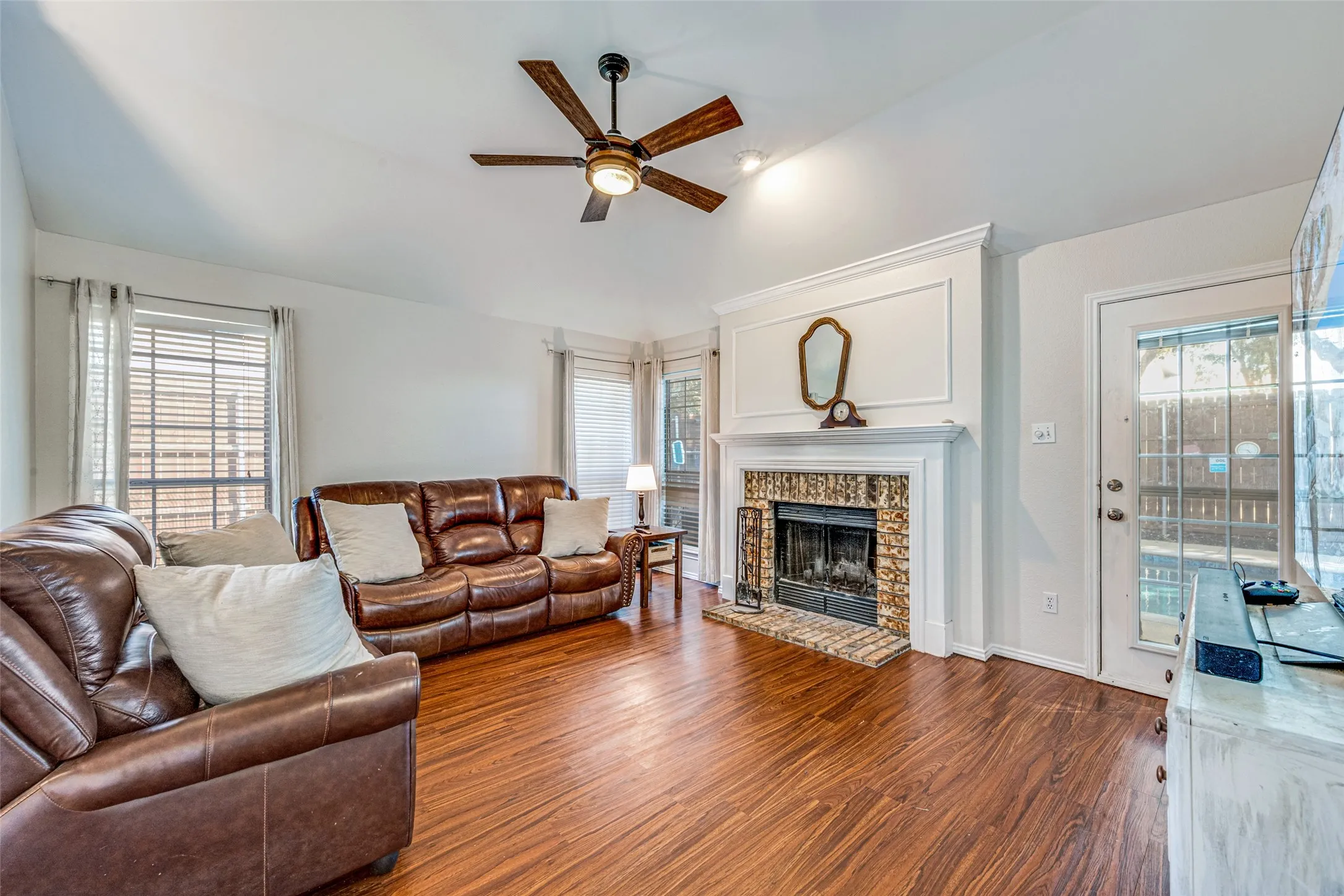 Vaulted ceiling with a wood-finish ceiling fan, wood-finish flooring, brick-surround fireplace with white mantel, glass-paned exterior door, and white window blinds
