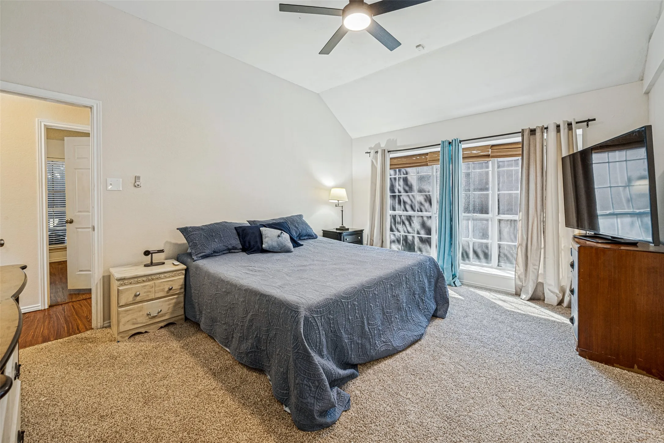 Vaulted ceiling, ceiling fan with integrated lighting, two windows with blinds, carpeted flooring, and a doorway with wood-finish flooring visible beyond
