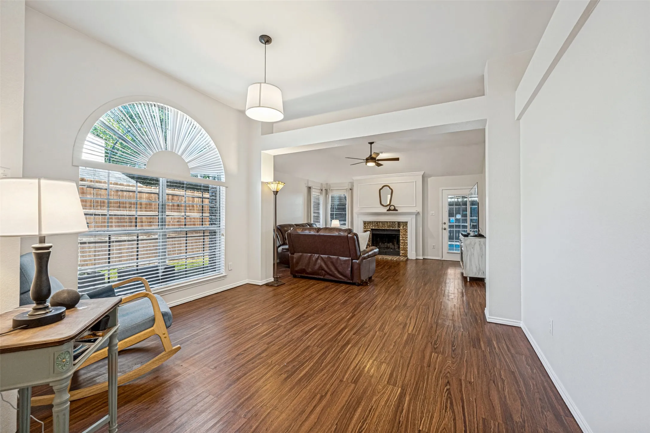 Wood-finish flooring throughout, arched window with horizontal blinds, overhead pendant lighting, and a fireplace with a white mantel and brick surround