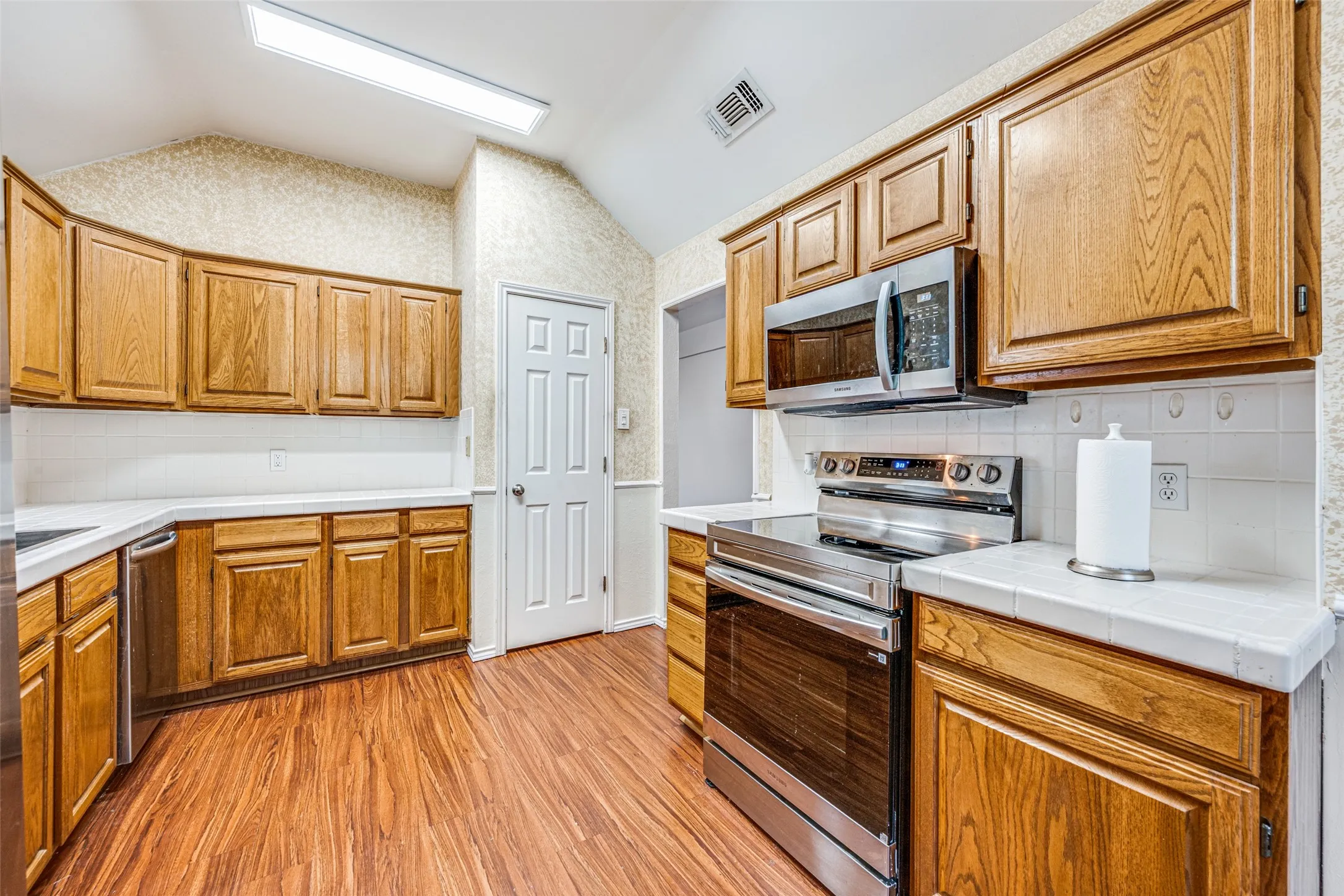Wood-finish flooring, wood cabinetry, white tile countertops, stainless steel appliances, and a vaulted ceiling with recessed lighting