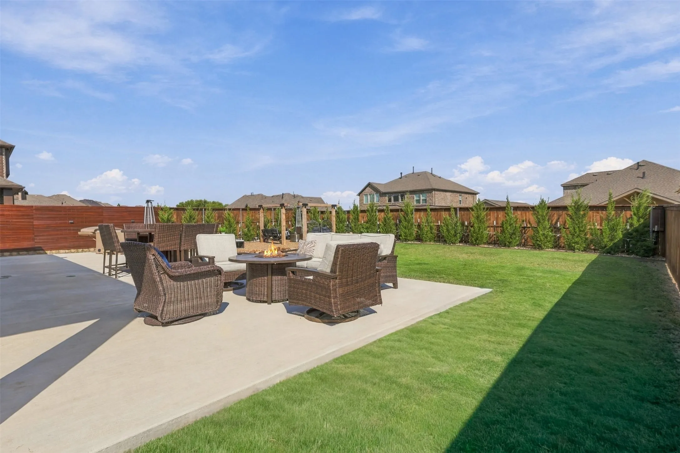 Expansive concrete patio with a fire pit feature, surrounded by a manicured lawn and a privacy fence with mature plantings