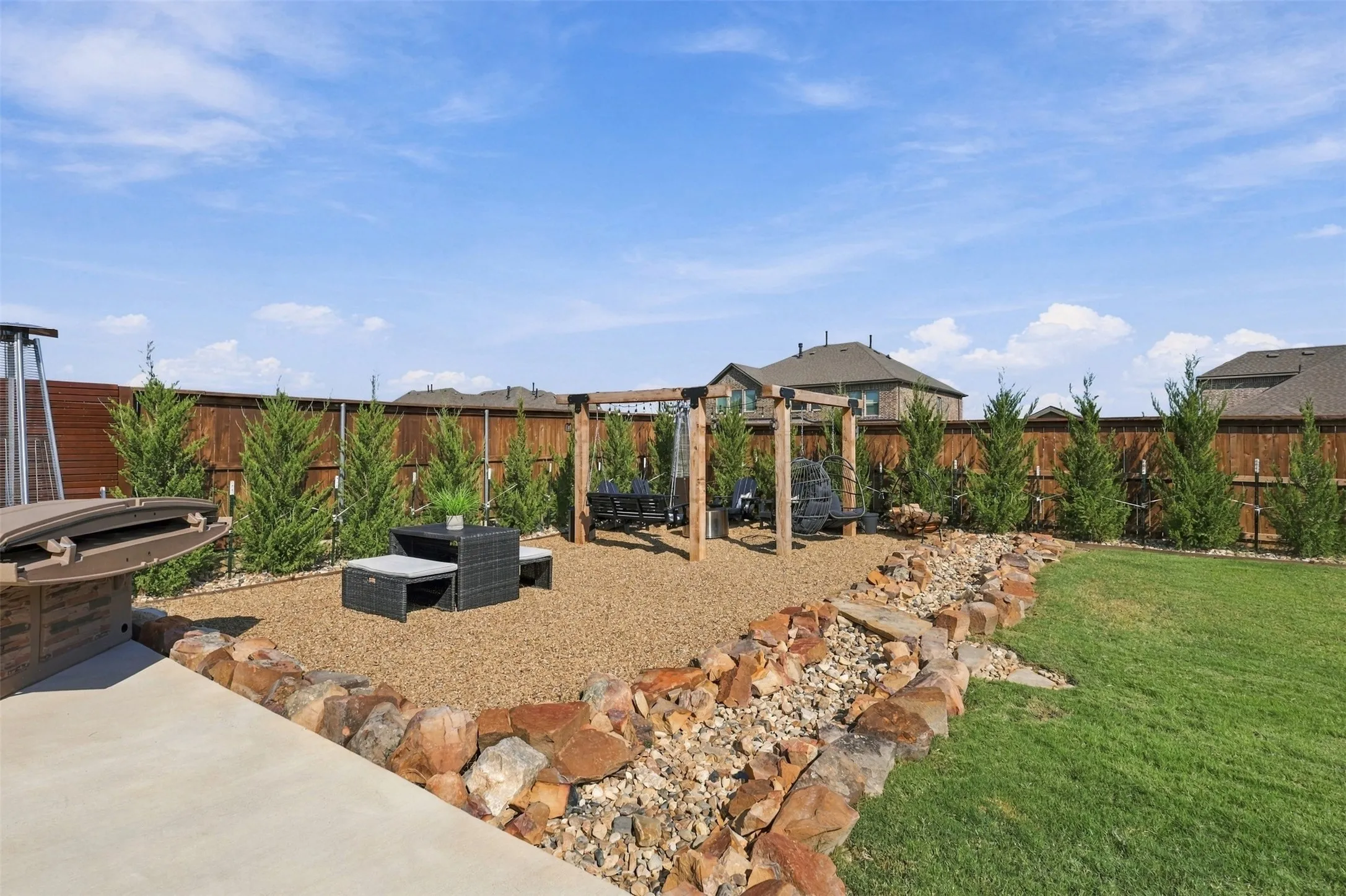Expansive backyard featuring a manicured lawn, a gravel patio area, a wooden privacy fence, and dry river bed with privacy trees