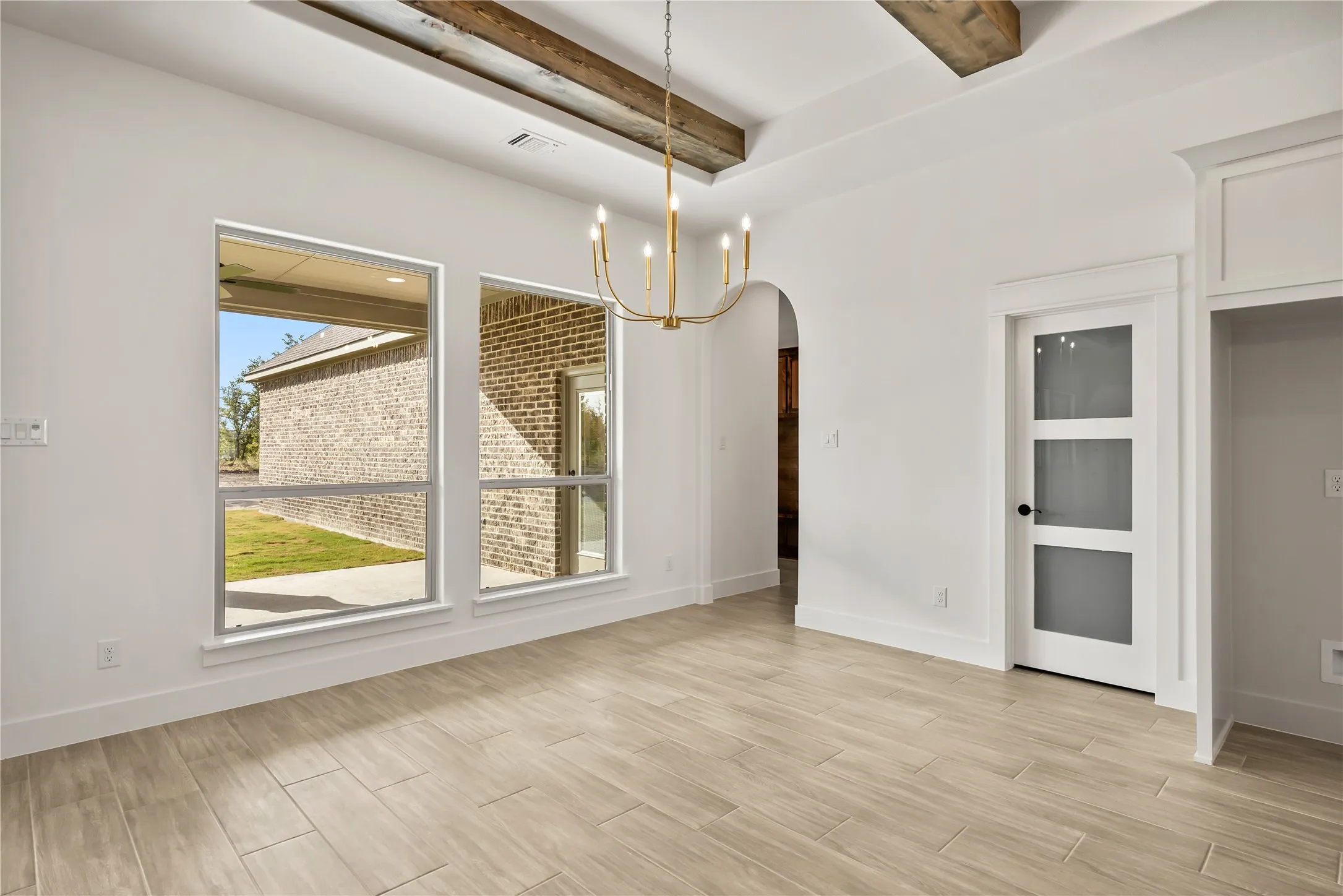 Recessed ceiling with exposed wood beams, wood-finish tile flooring, modern chandelier, arched doorway, and a white interior door with frosted glass panels