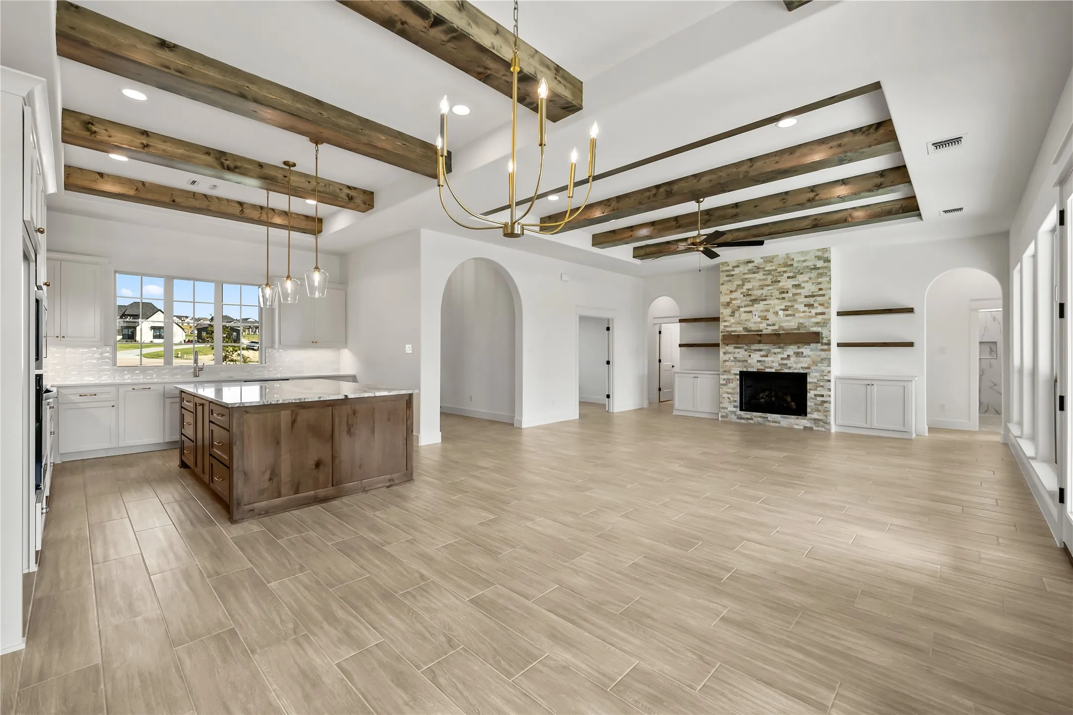 Vaulted ceiling with exposed wood beams, large windows, wood-finish flooring, stone-clad fireplace, and a kitchen island with stone countertop