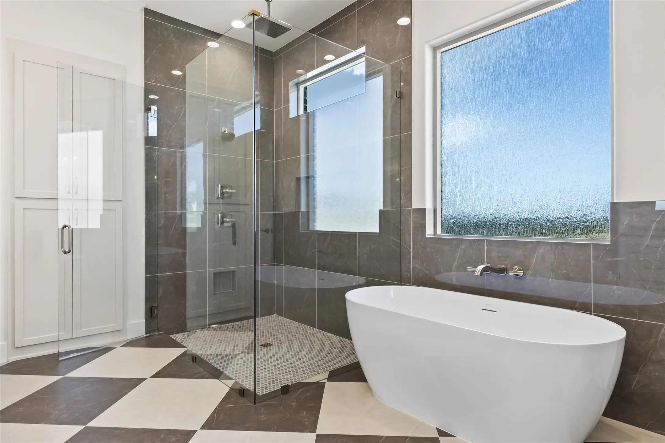 Freestanding soaking tub, glass-enclosed shower, built-in cabinetry, large privacy window, and checkered floor tile