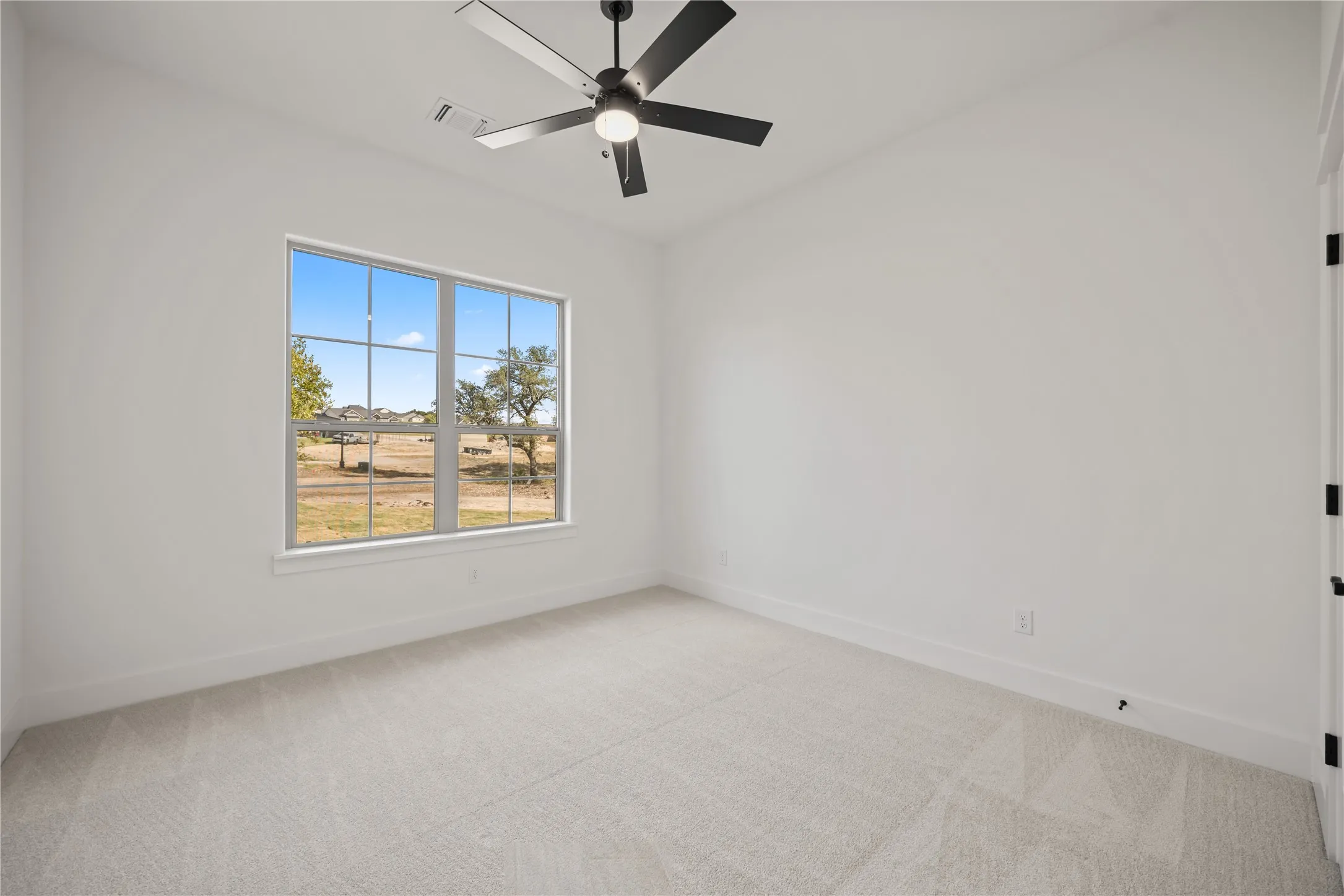 Spacious room with plush carpeting, a large window, a modern ceiling fan with lighting, and crisp white walls