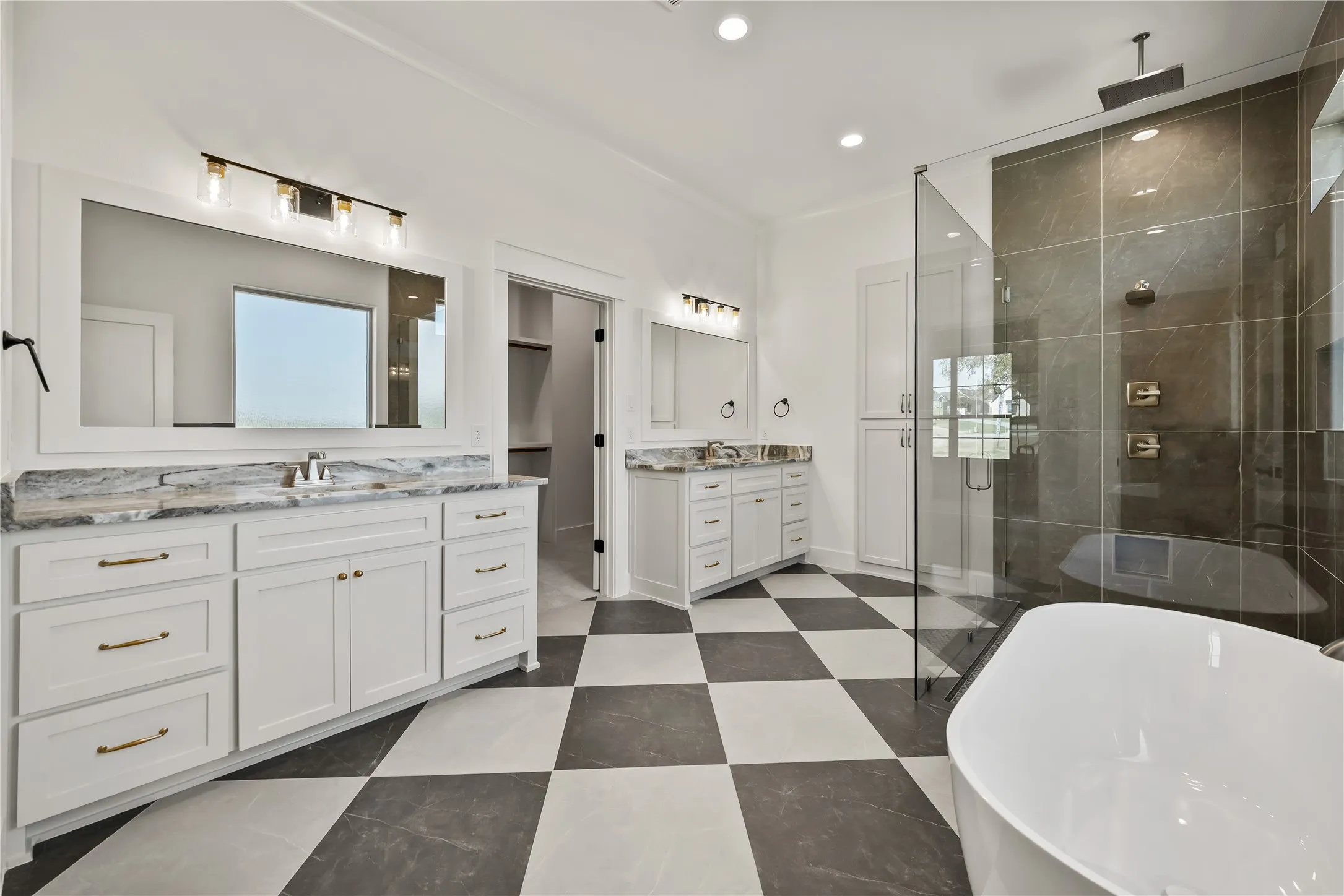 Dual vanities with stone countertops, a freestanding soaking tub, a glass-enclosed shower with dark tile surround, checkerboard tile flooring, and built-in linen cabinetry