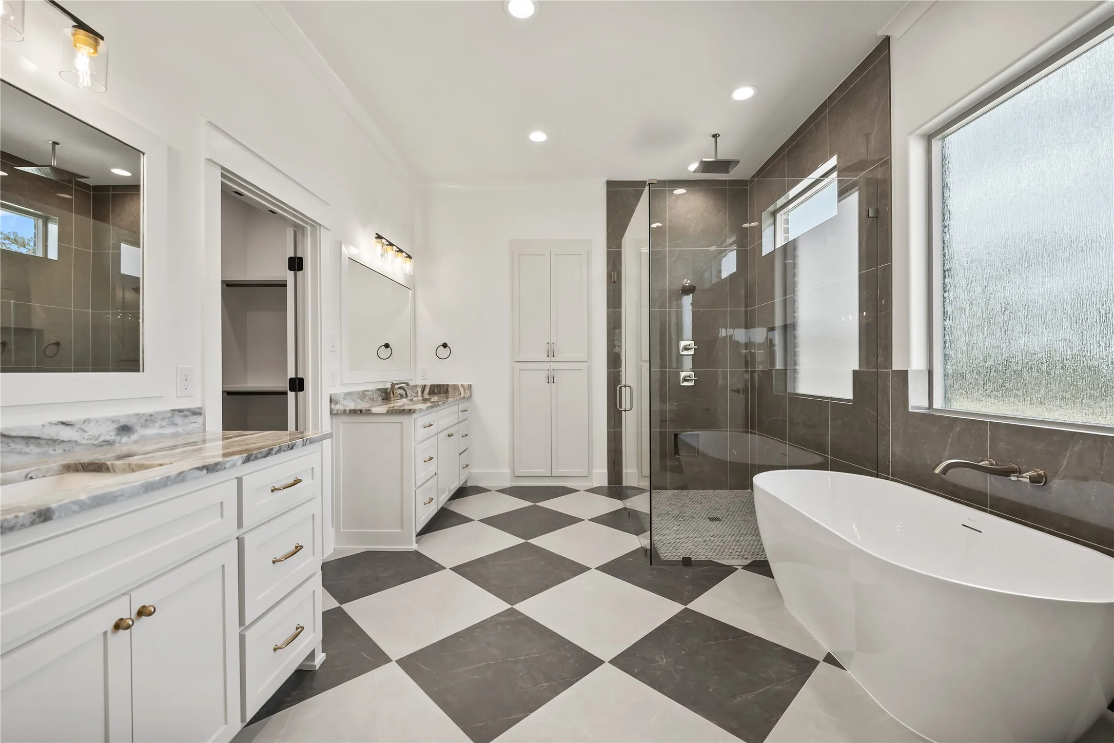 Freestanding soaking tub, frameless glass shower enclosure with bench, dual vanity sinks with stone countertops, checkered tile flooring, and built-in storage cabinetry