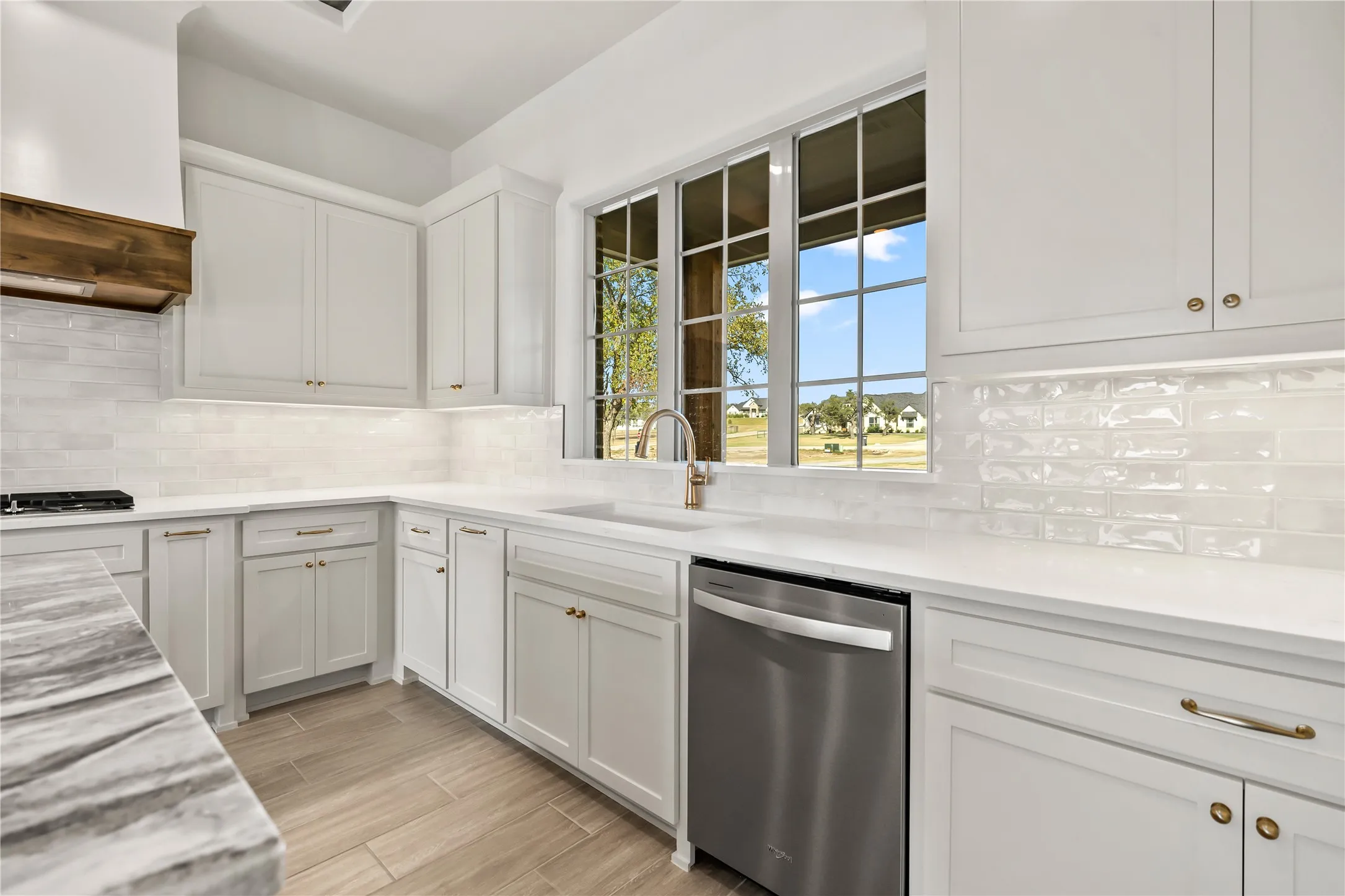White cabinetry with gold-tone hardware, a stainless steel dishwasher, a single basin sink with a gold-tone faucet, wood-finish flooring, and a white subway tile backsplash