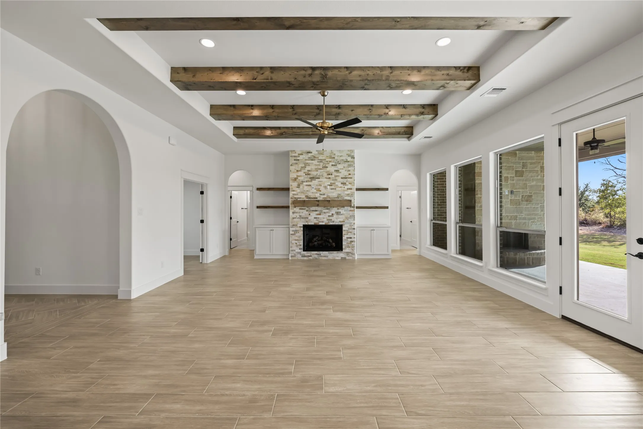 Vaulted ceiling with exposed wood beams, stone-clad fireplace with wood mantle, built-in cabinetry with open shelving, large windows, and a glass-paneled door