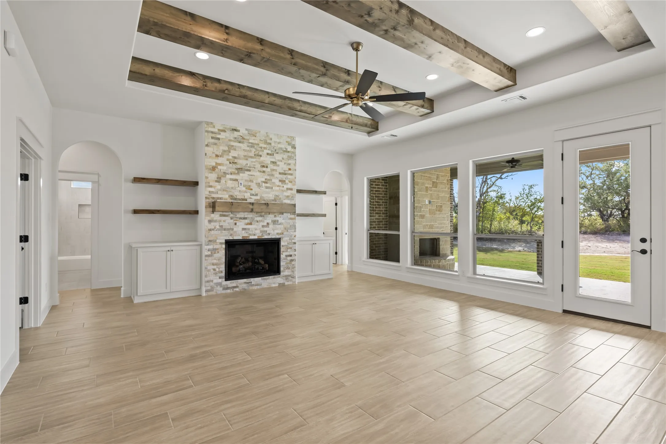Tray ceiling with exposed wood beams, stone-clad fireplace with wood mantel, built-in cabinetry with open shelving, large format tile flooring, and multiple exterior access points