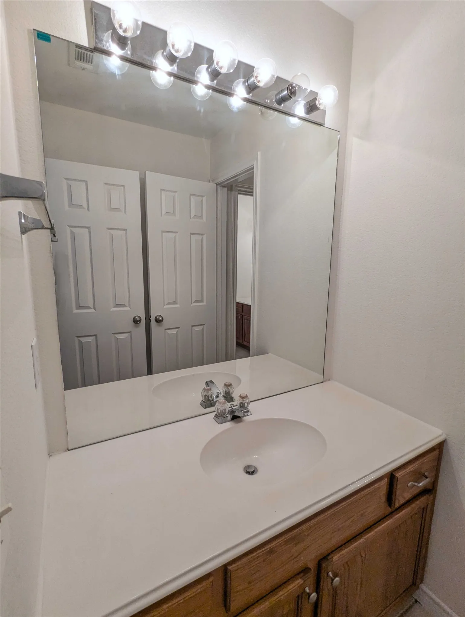 Vanity with a white countertop and integrated sink, chrome faucet, wood-finish cabinetry with silver-toned hardware, and a large wall-mounted mirror