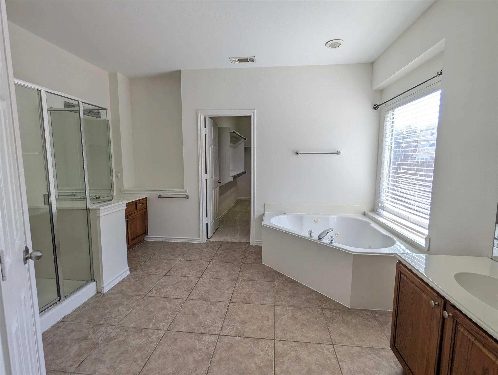 Tiled flooring throughout, corner jetted tub, glass-enclosed shower, wood-finish vanity with an integrated sink, and recessed lighting