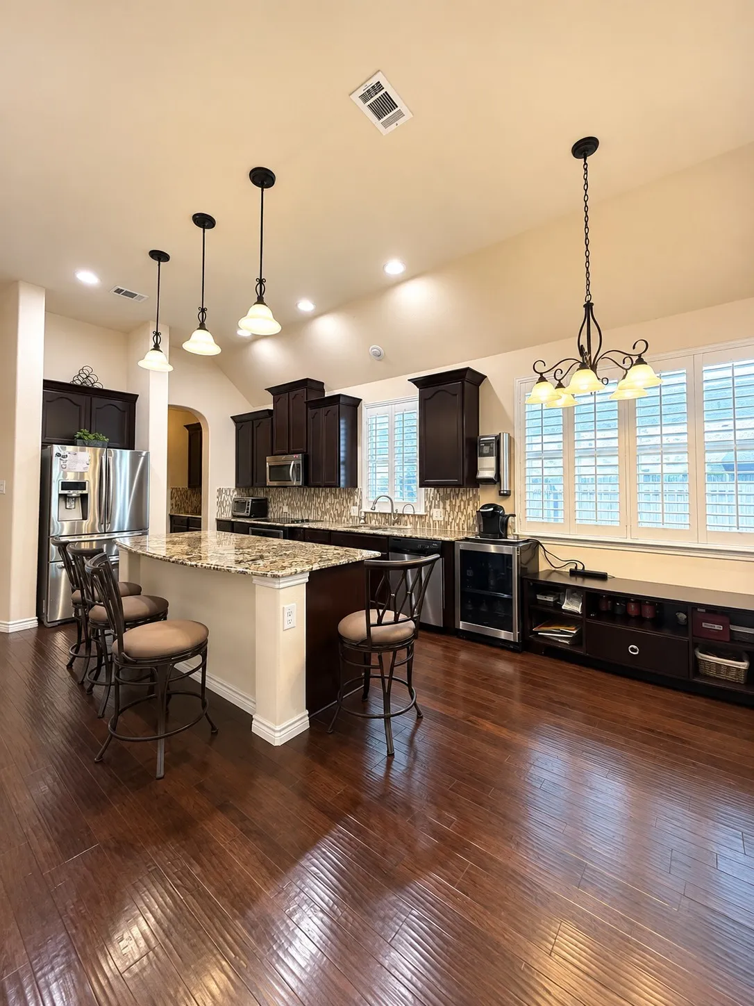 Spacious kitchen featuring dark wood cabinetry, granite countertops, wood-finish flooring, stainless steel appliances, and a central island with pendant lighting