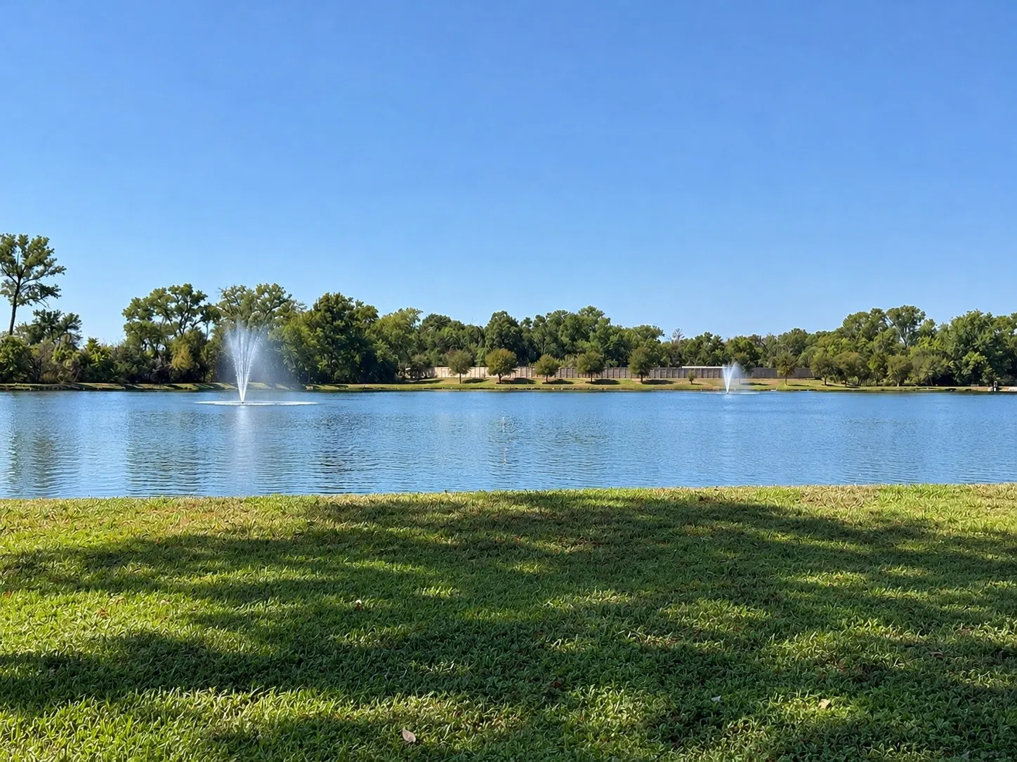 Expansive body of water with multiple fountains, bordered by lush green landscaping and mature trees
