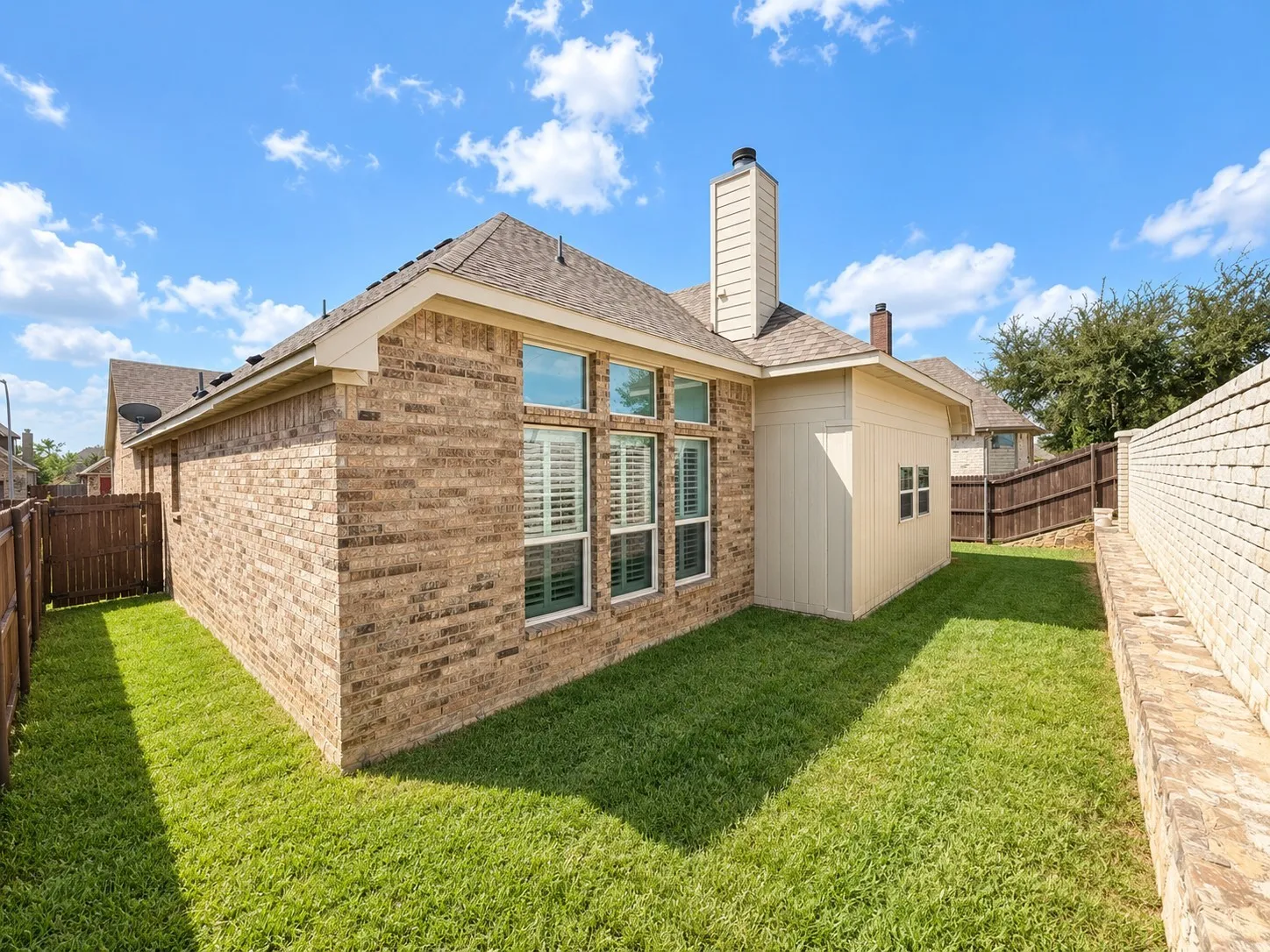 Brick exterior with multiple windows featuring plantation shutters, a shingled roof, a chimney, and a wooden privacy fence