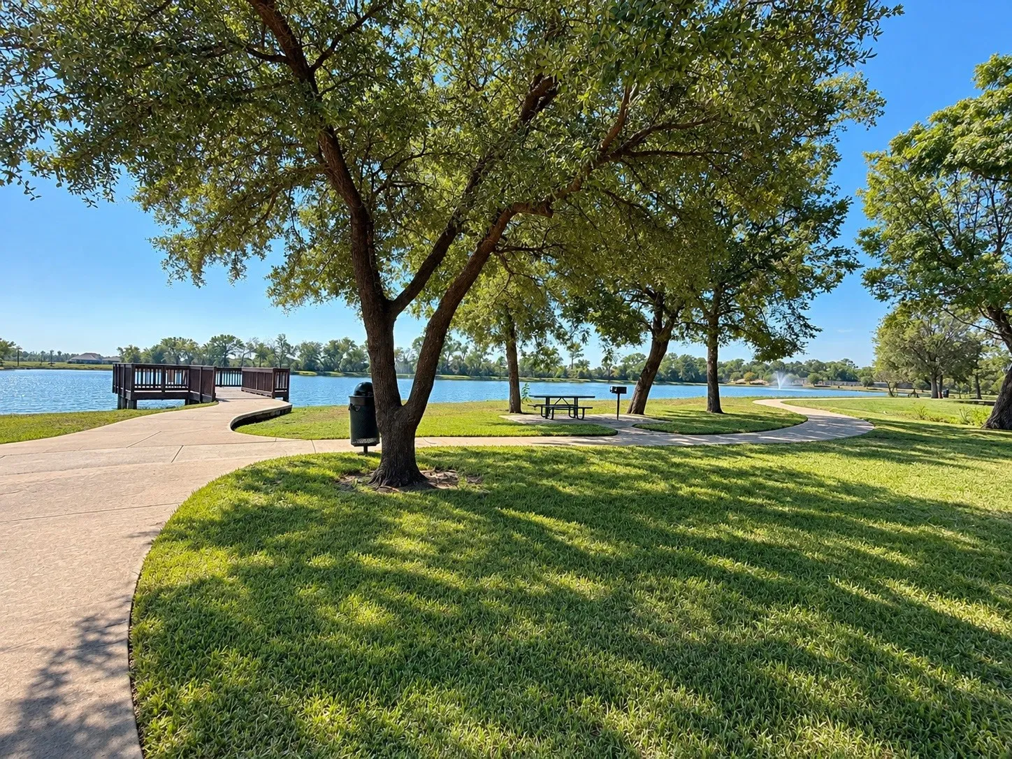 Expansive green space with mature trees, a concrete pathway, and a wooden pier extending over the water