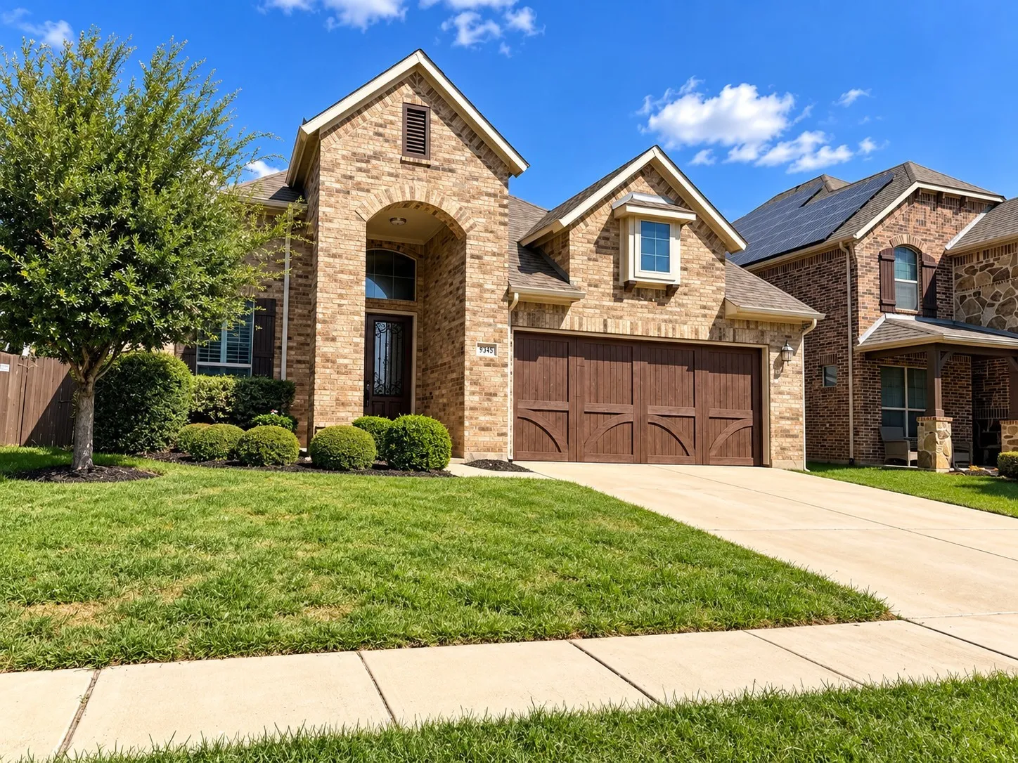 Brick façade with an arched entryway, mature landscaping, an attached two-car garage with wood-finish doors, and a concrete driveway