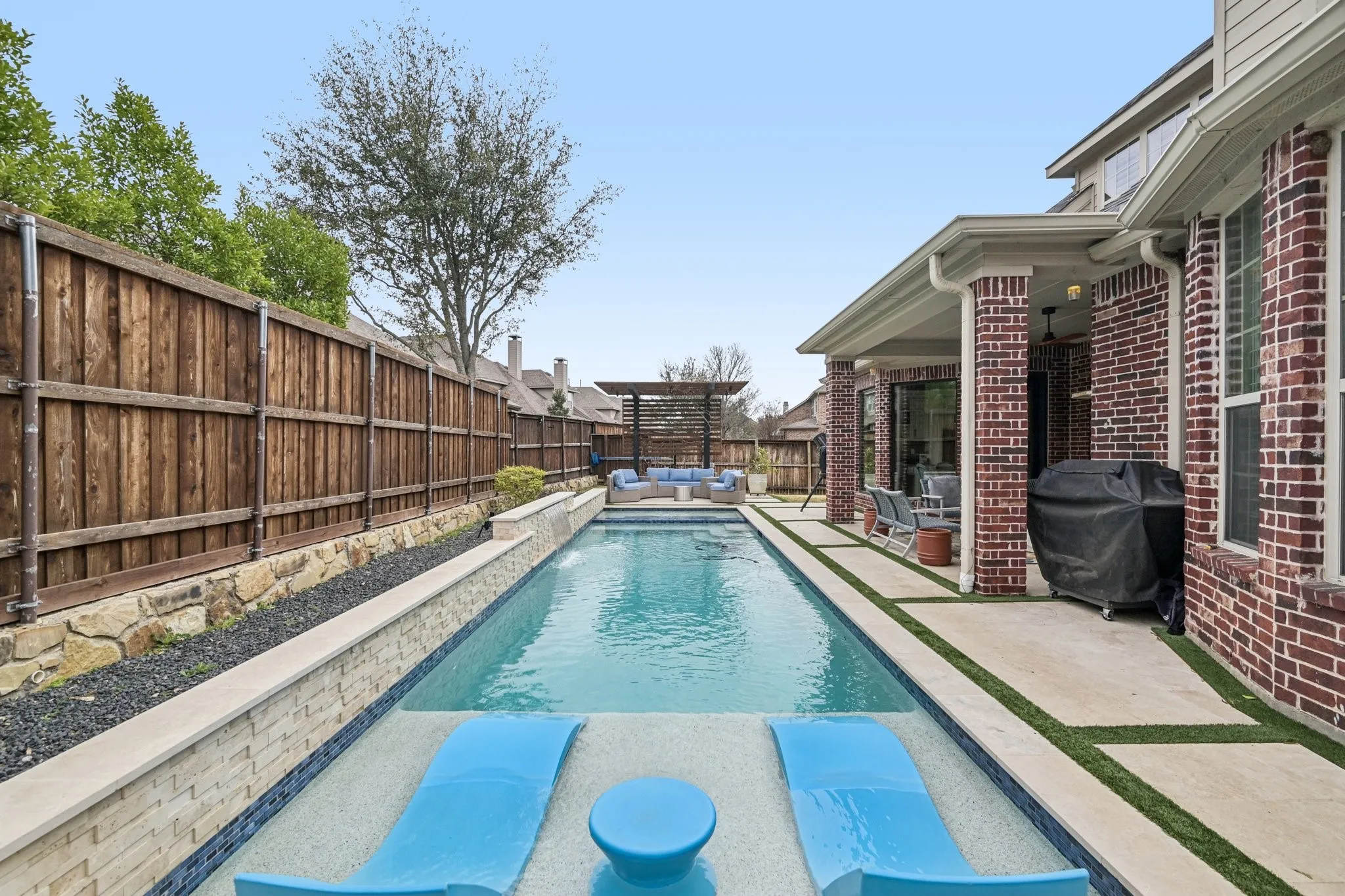 Rectangular swimming pool featuring integrated lounge seating, a stone retaining wall with dark rock ground cover, and a wood privacy fence