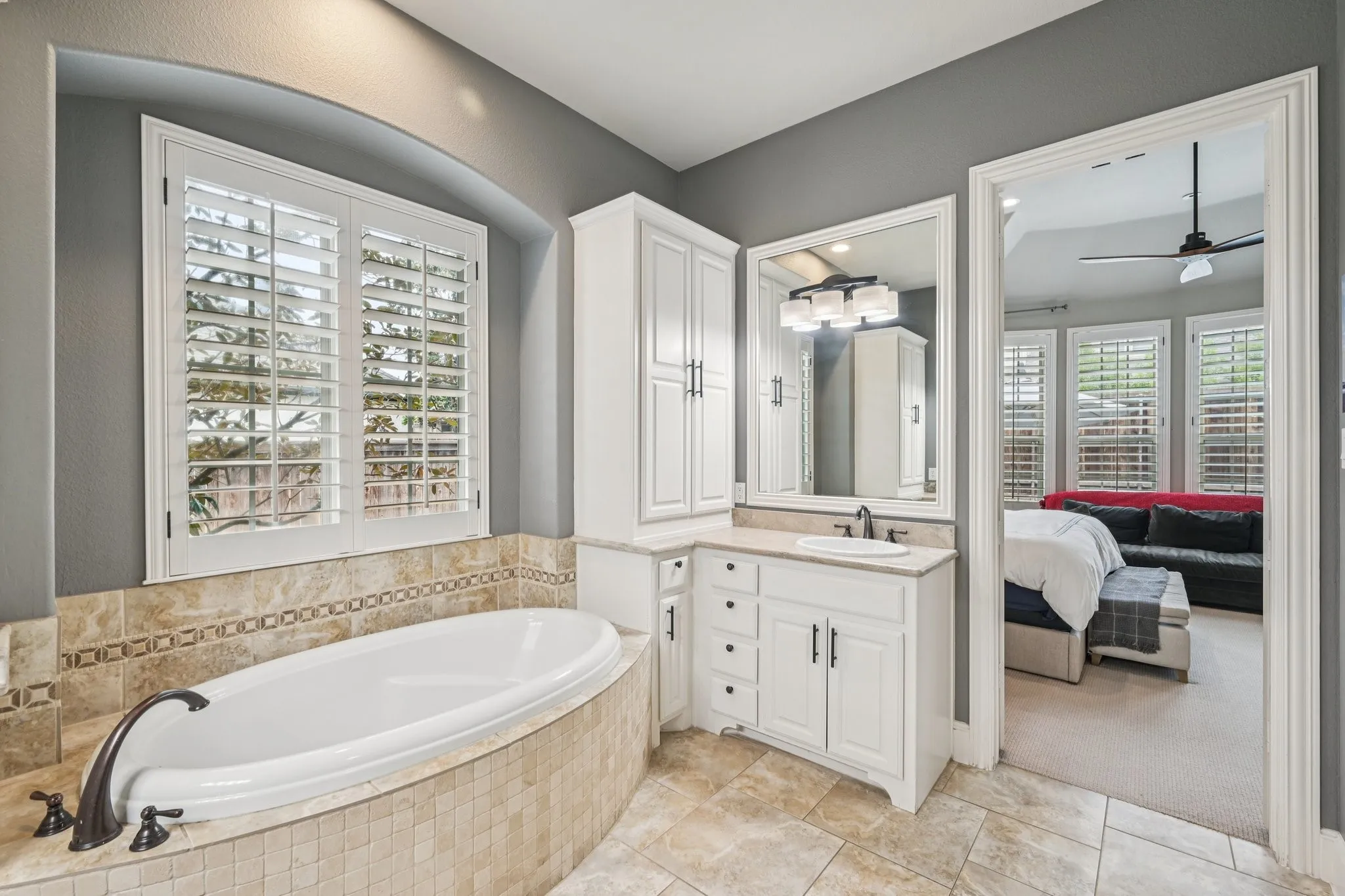 Oval soaking tub with tiled surround, white plantation shutters, white vanity with integrated sink, and tiled flooring