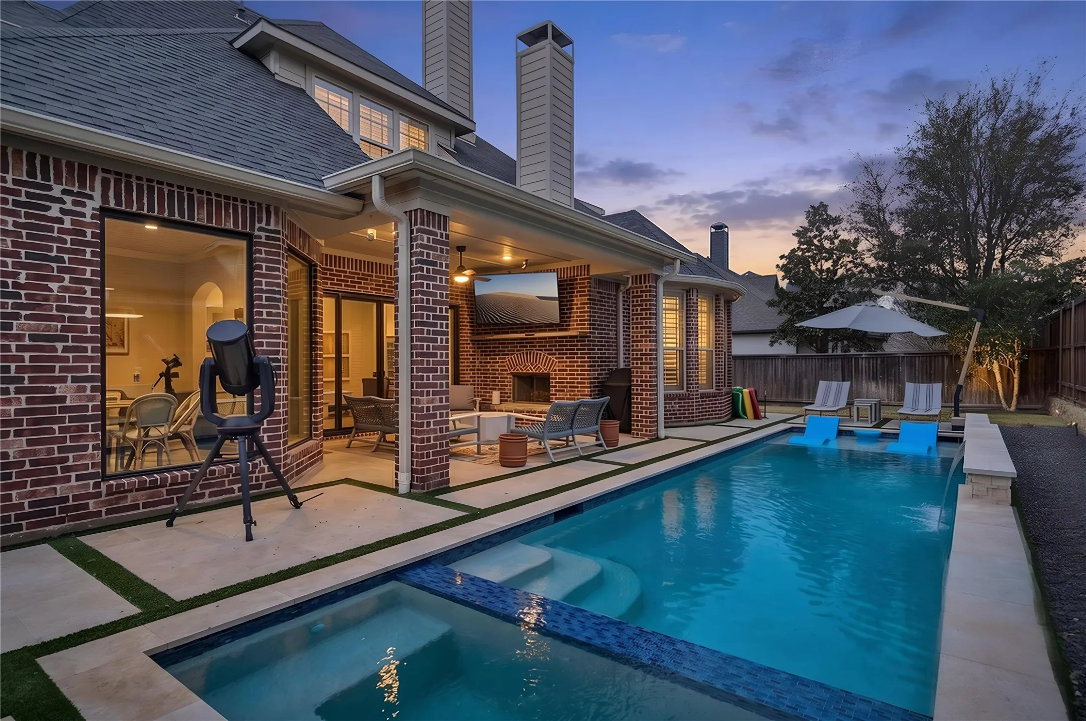 Brick exterior with multiple windows, covered patio, outdoor fireplace, built-in television, and an integrated pool and spa