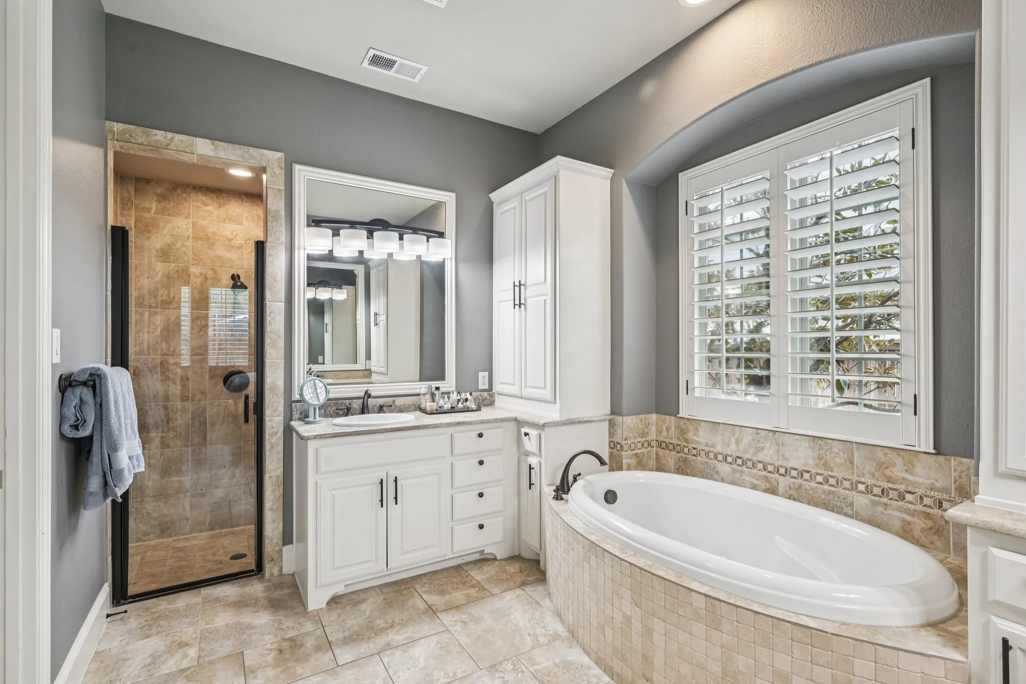 Tiled walk-in shower with glass enclosure, single vanity with countertop, built-in soaking tub with tiled surround, white built-in cabinetry, and window with plantation shutters