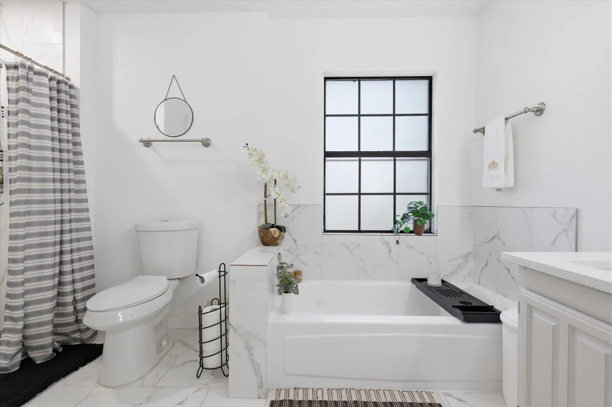 White subway tile shower surround, integrated bathtub, black grid window, white vanity cabinetry, and white tile flooring