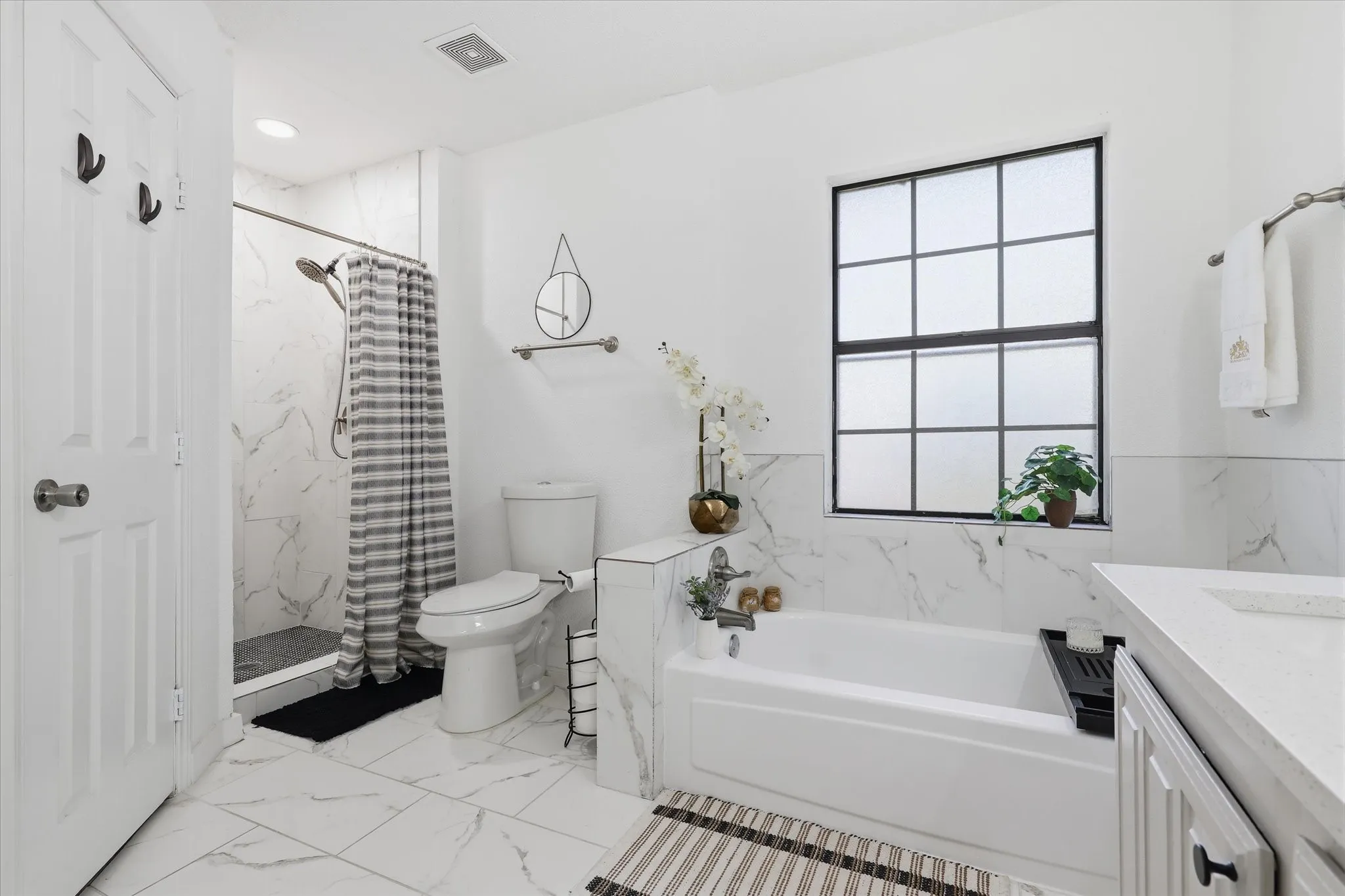 White tiled flooring, white wall tiling, separate shower enclosure with glass panel, white bathtub, and a window with black grid panes