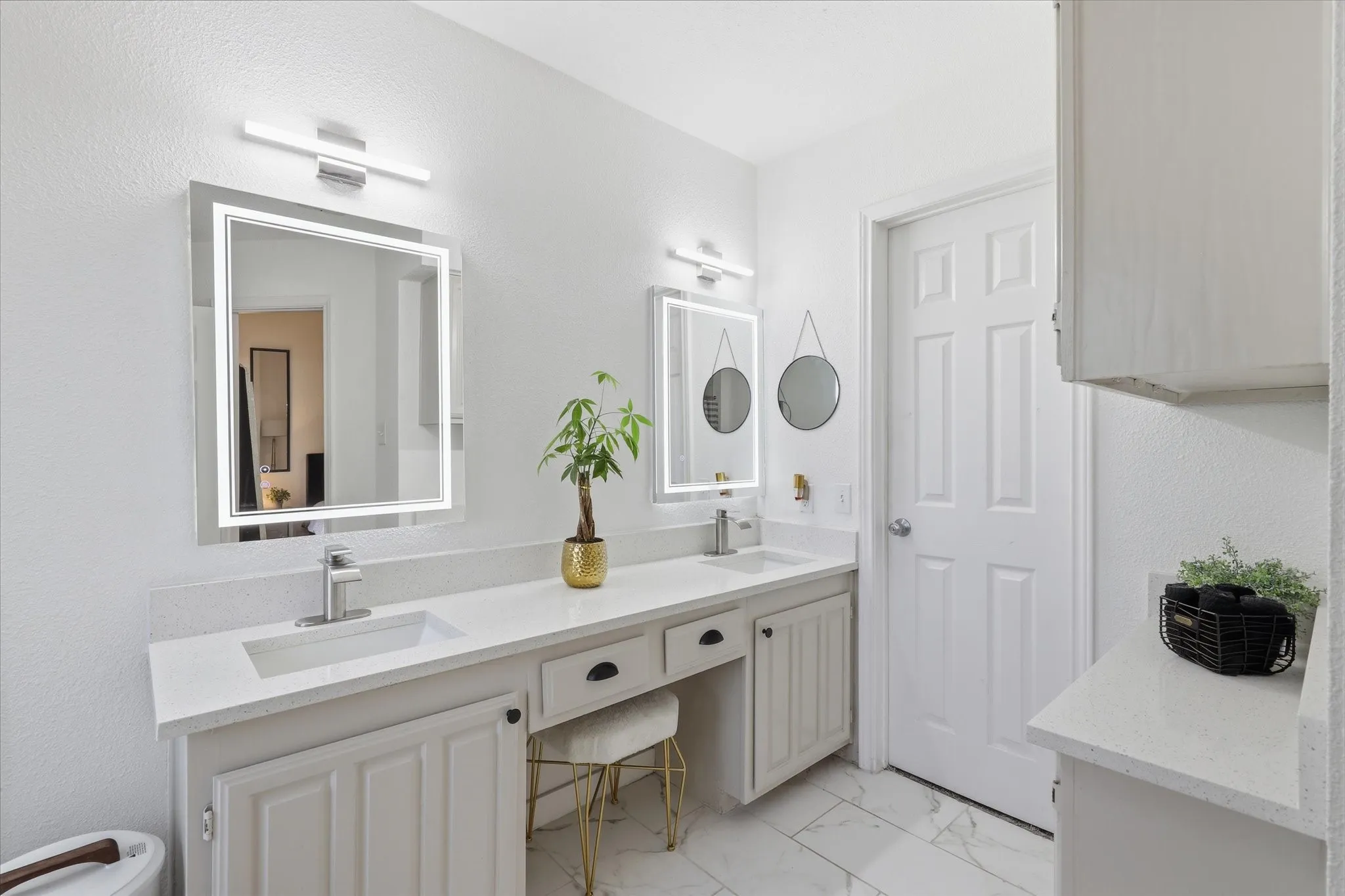 Dual vanities with integrated sinks, illuminated mirrors, polished chrome fixtures, white paneled cabinetry, and white tile flooring