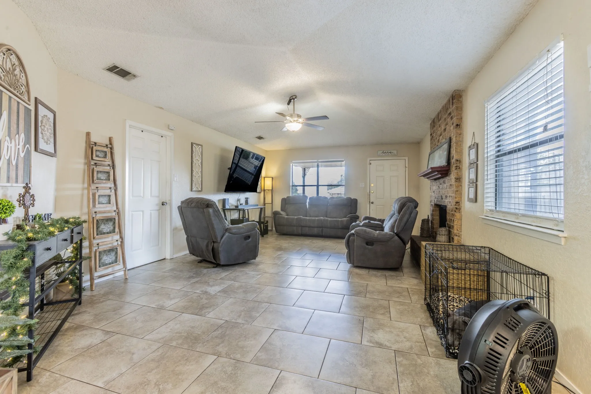 Spacious living area featuring tile flooring, a ceiling fan with integrated lighting, multiple windows, and a brick-clad fireplace