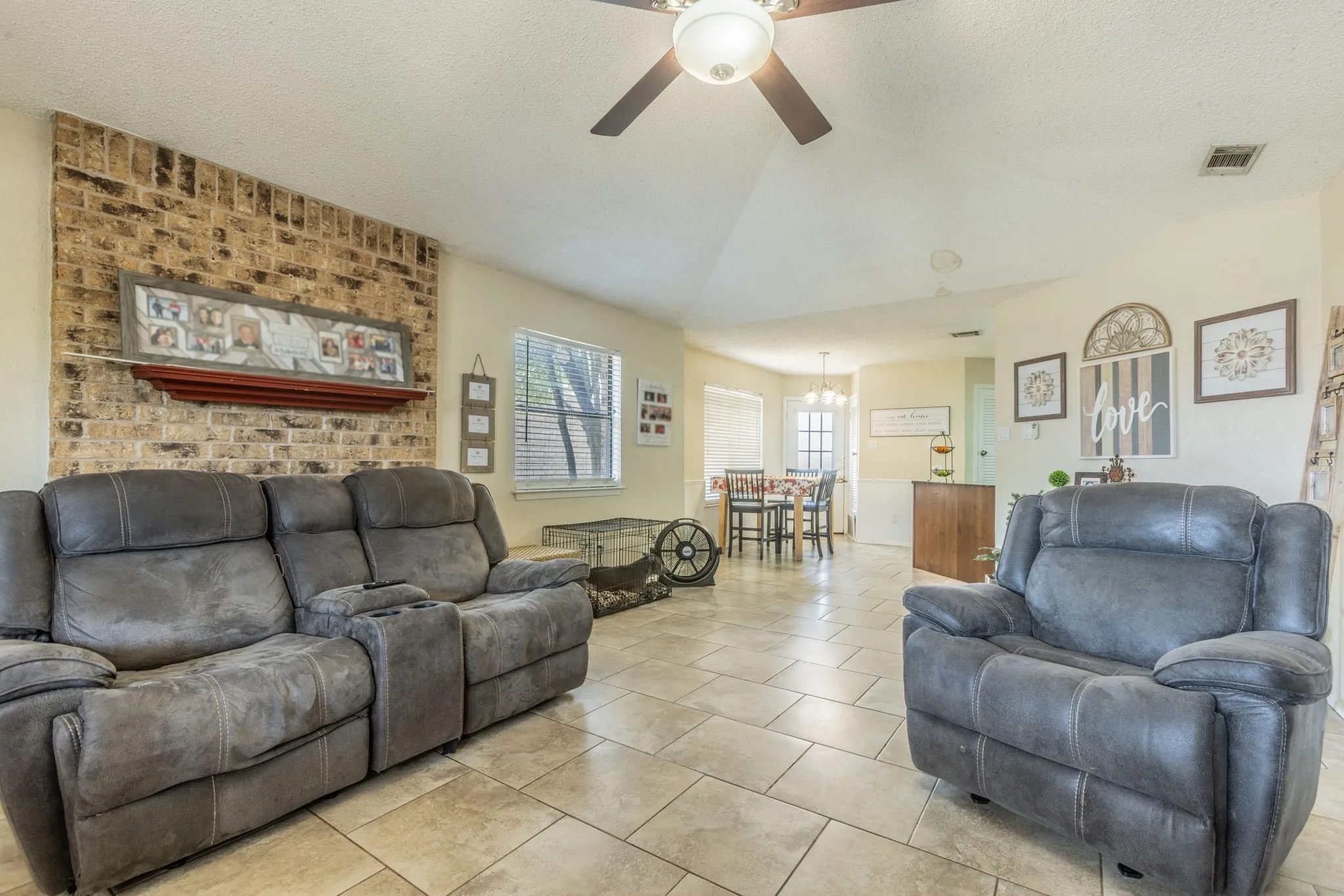 Vaulted ceiling with a ceiling fan, brick accent wall with a mantel, tiled flooring, and a dining area with a chandelier