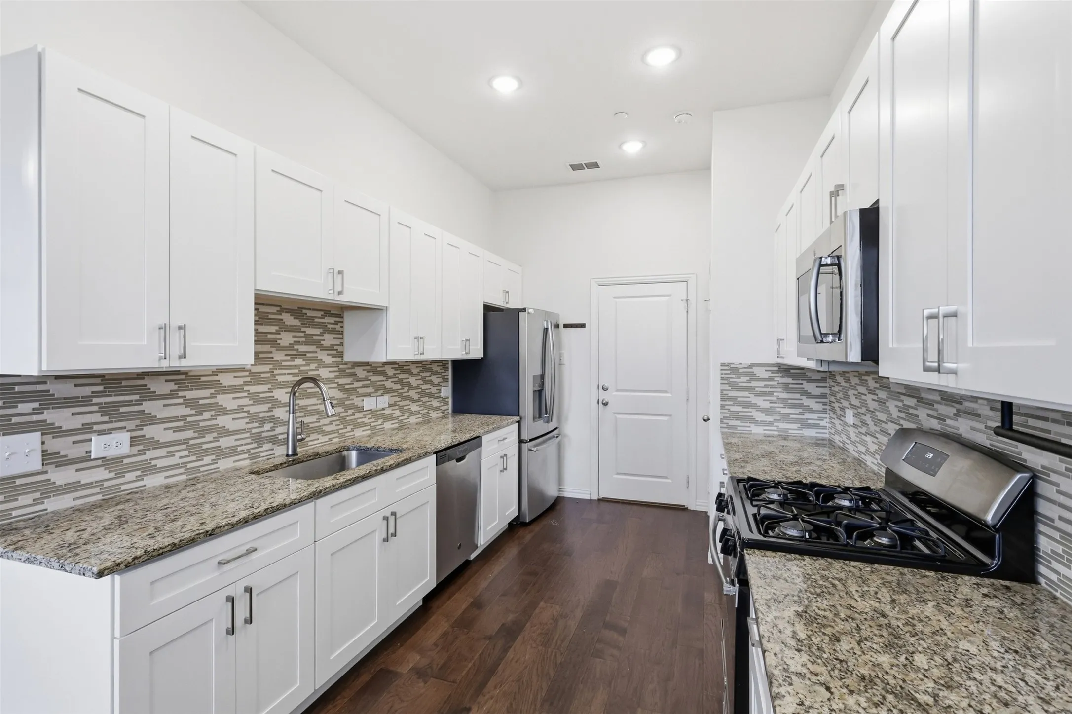 Kitchen featuring white cabinetry, light-toned granite countertops, a mosaic tile backsplash, stainless steel appliances, and dark wood-finish flooring