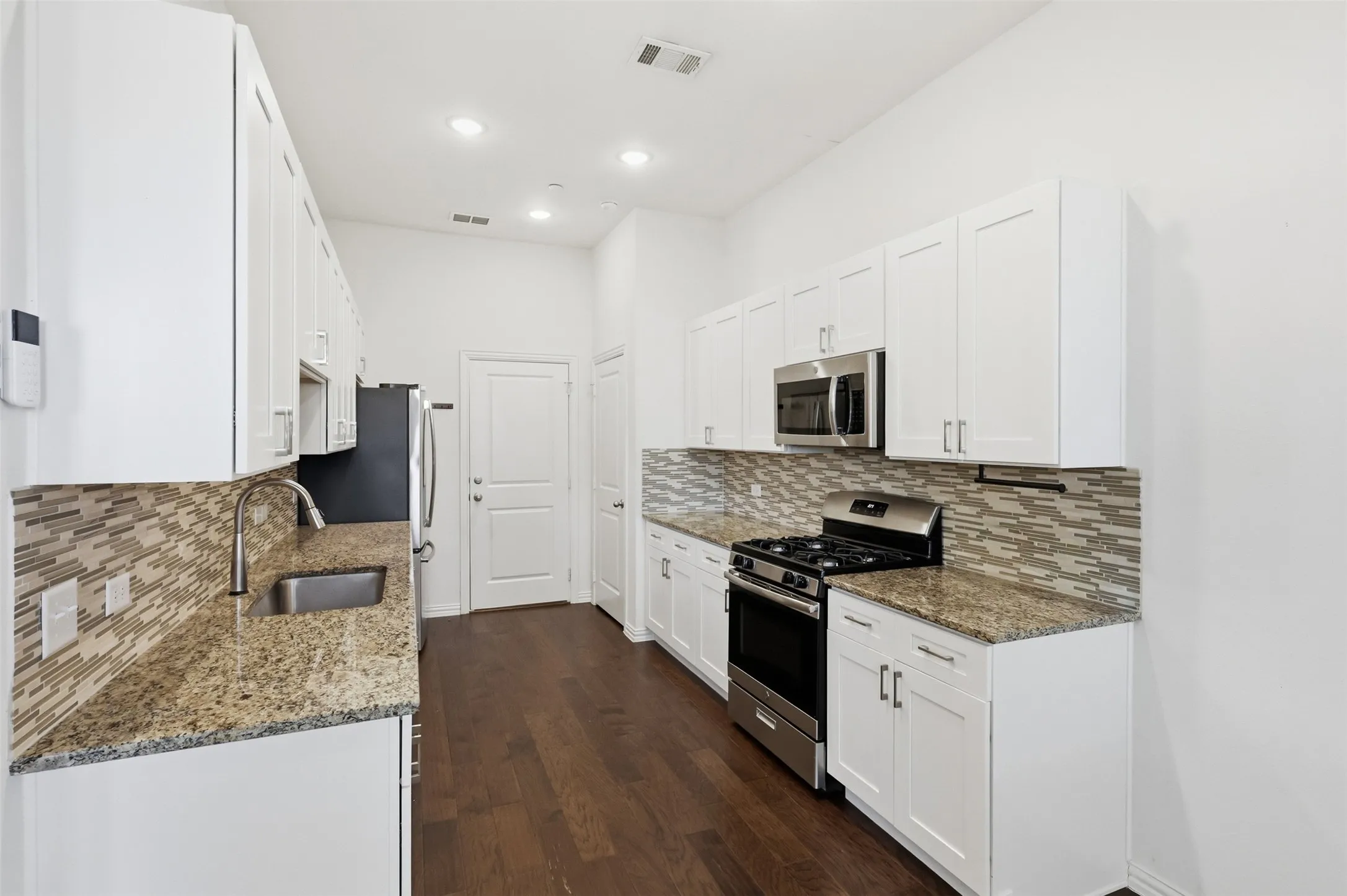 Galley kitchen featuring white cabinetry, stainless steel appliances, granite countertops, a mosaic tile backsplash, and dark wood-finish flooring