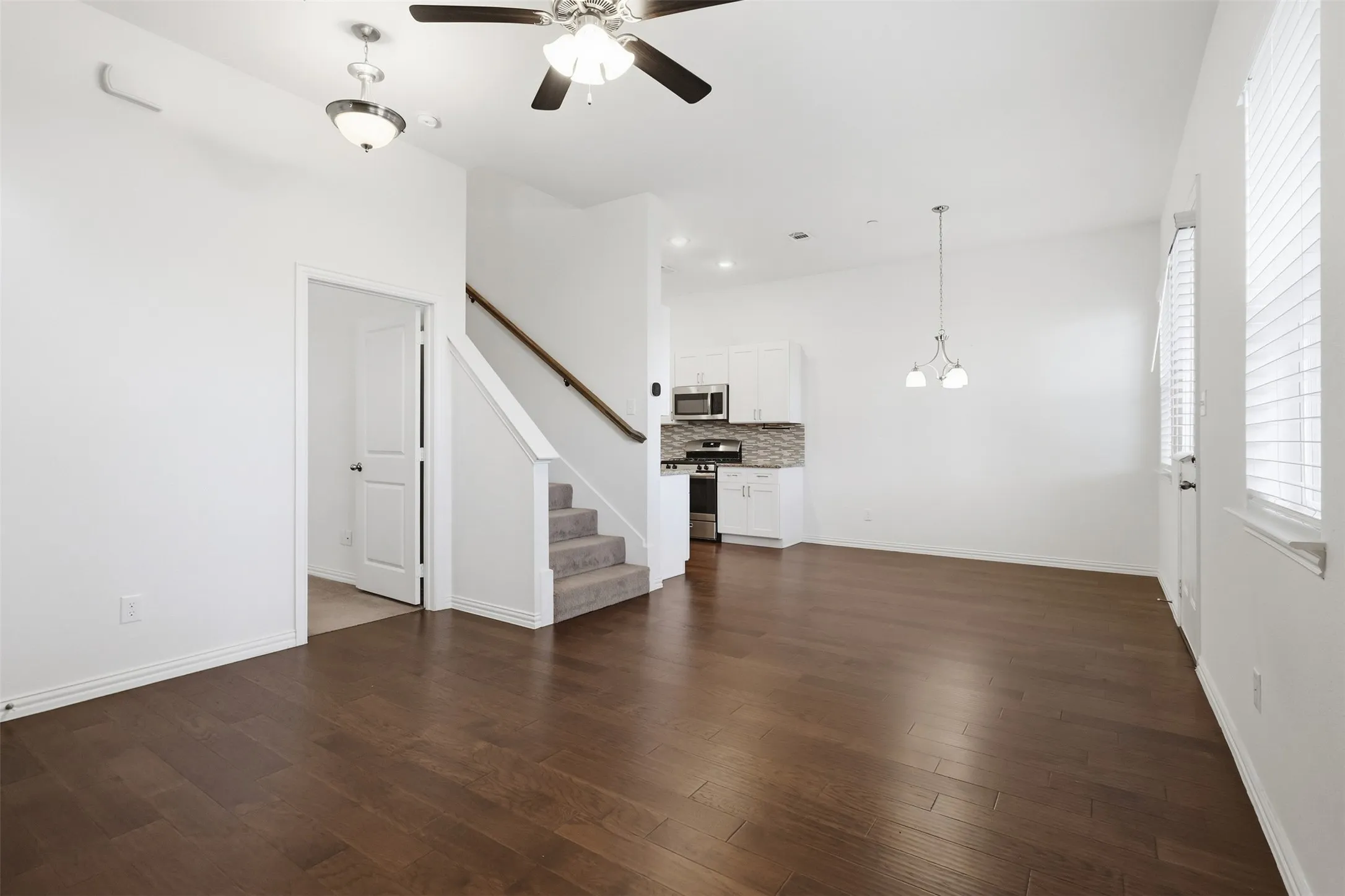 Wood-finish flooring, staircase with carpeted treads and wood handrail, ceiling fan, pendant lighting, and white cabinetry