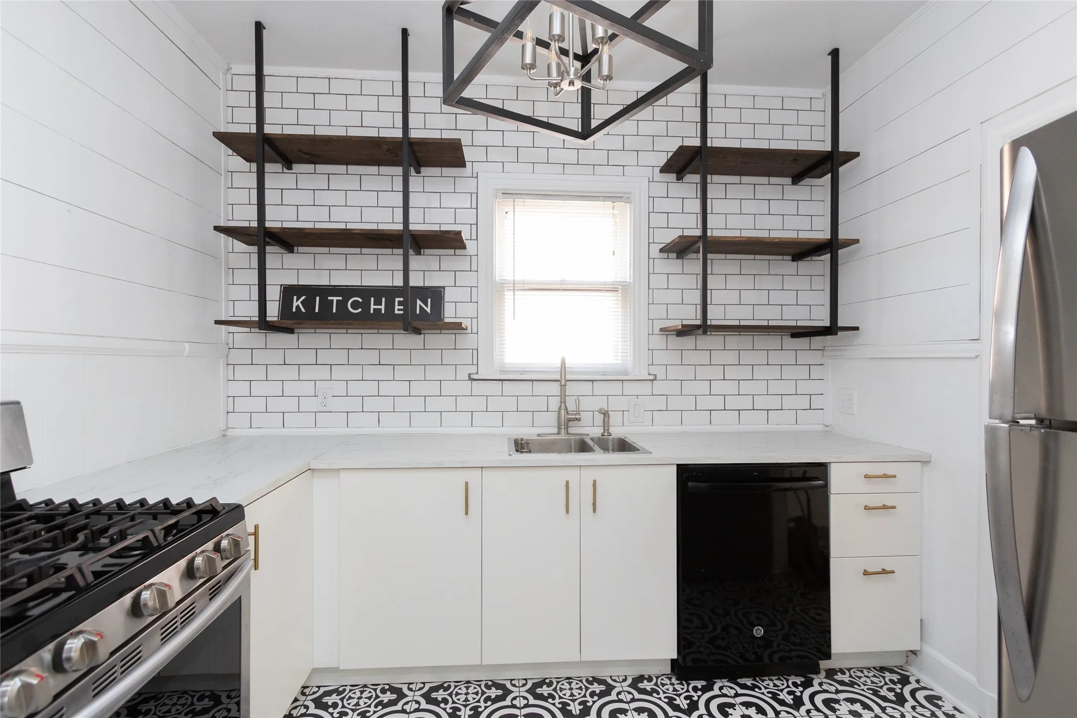 White subway tile backsplash, white shaker-style cabinetry with gold-toned hardware, stainless steel gas range, and black-and-white patterned flooring