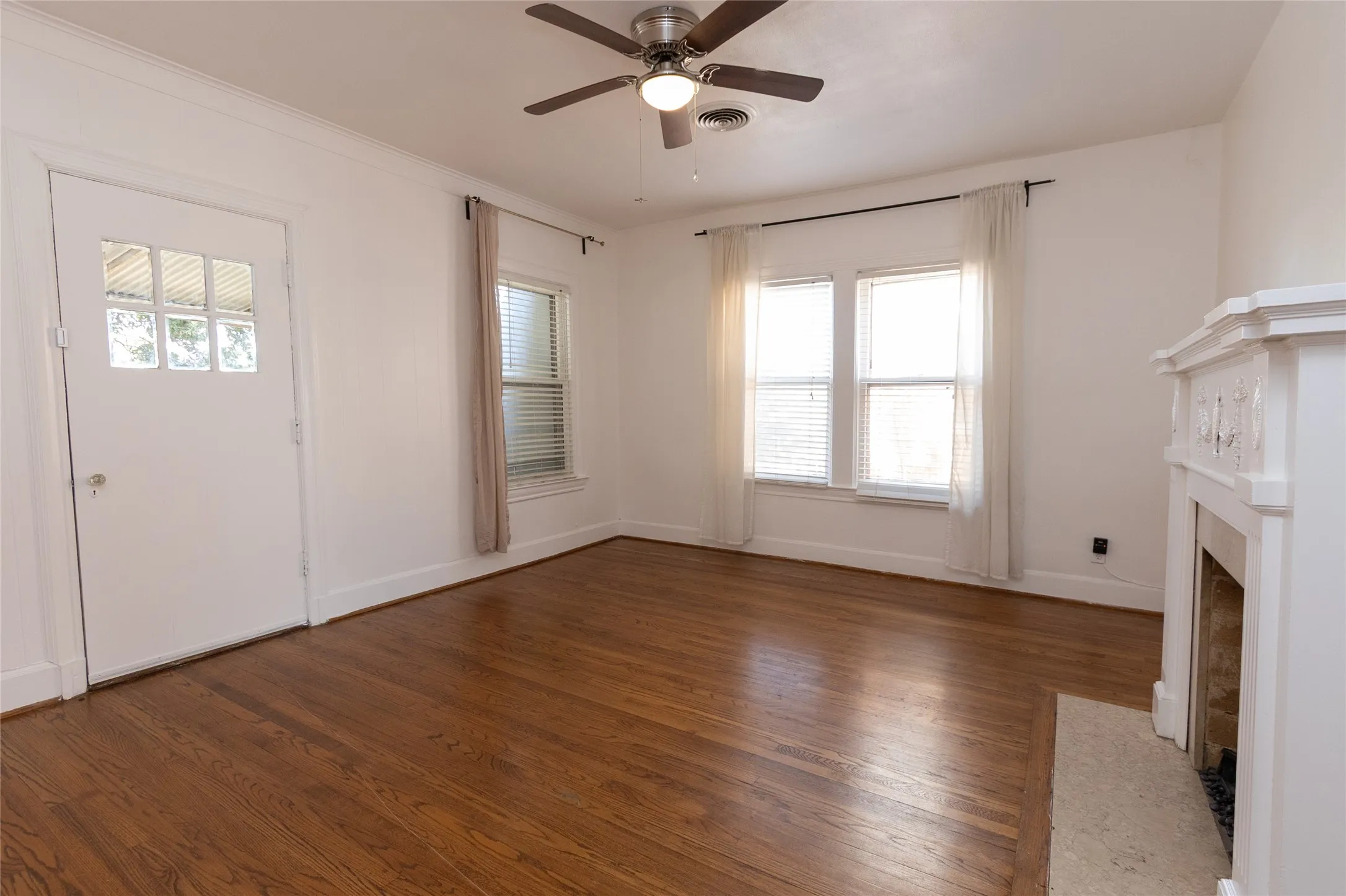 Hardwood flooring throughout, featuring a decorative fireplace with an ornate mantel, a ceiling fan with integrated lighting, and multiple windows for natural light