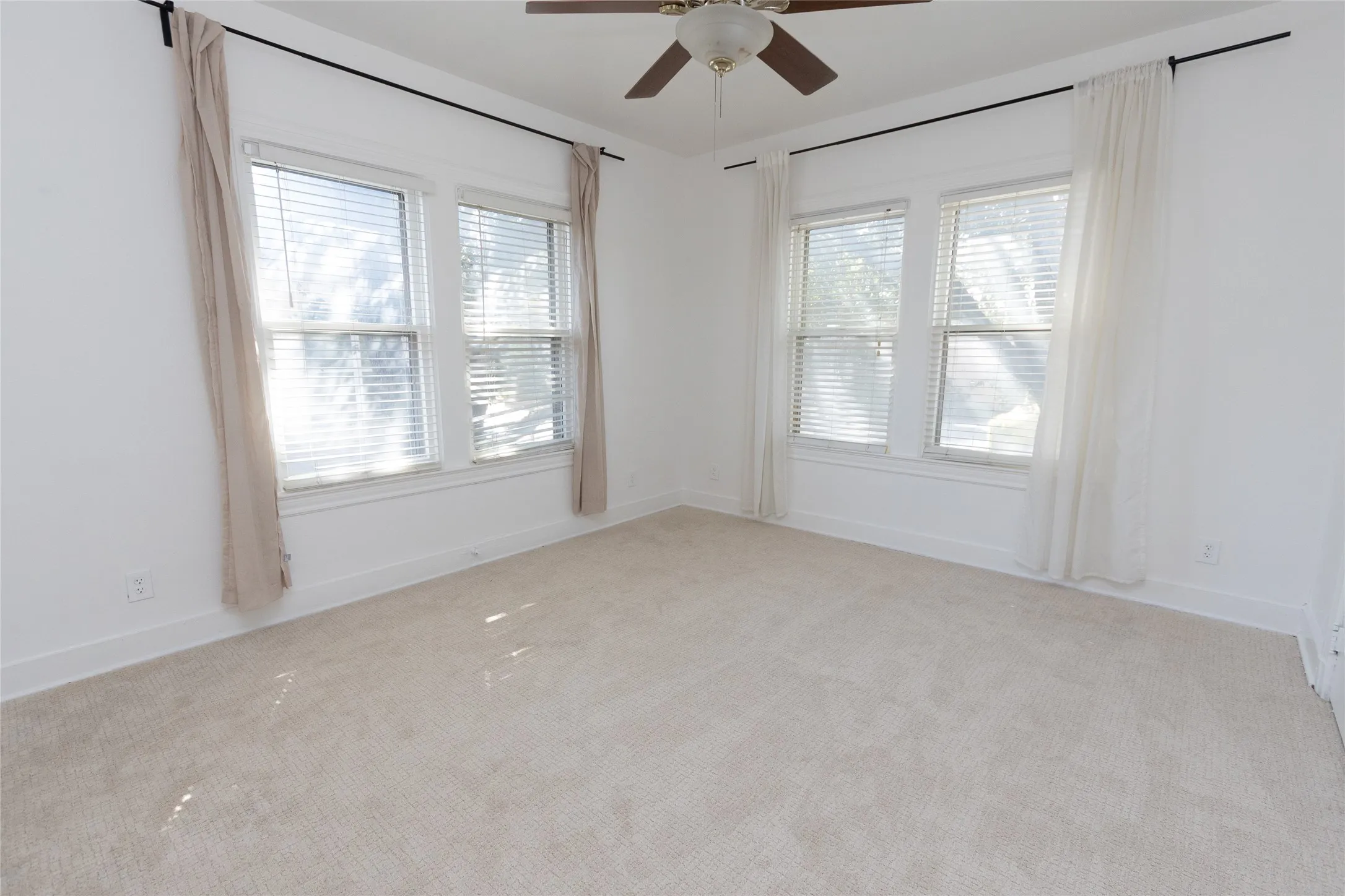 Expansive room featuring light-toned carpeting, multiple windows with blinds, a ceiling fan, and crisp white walls